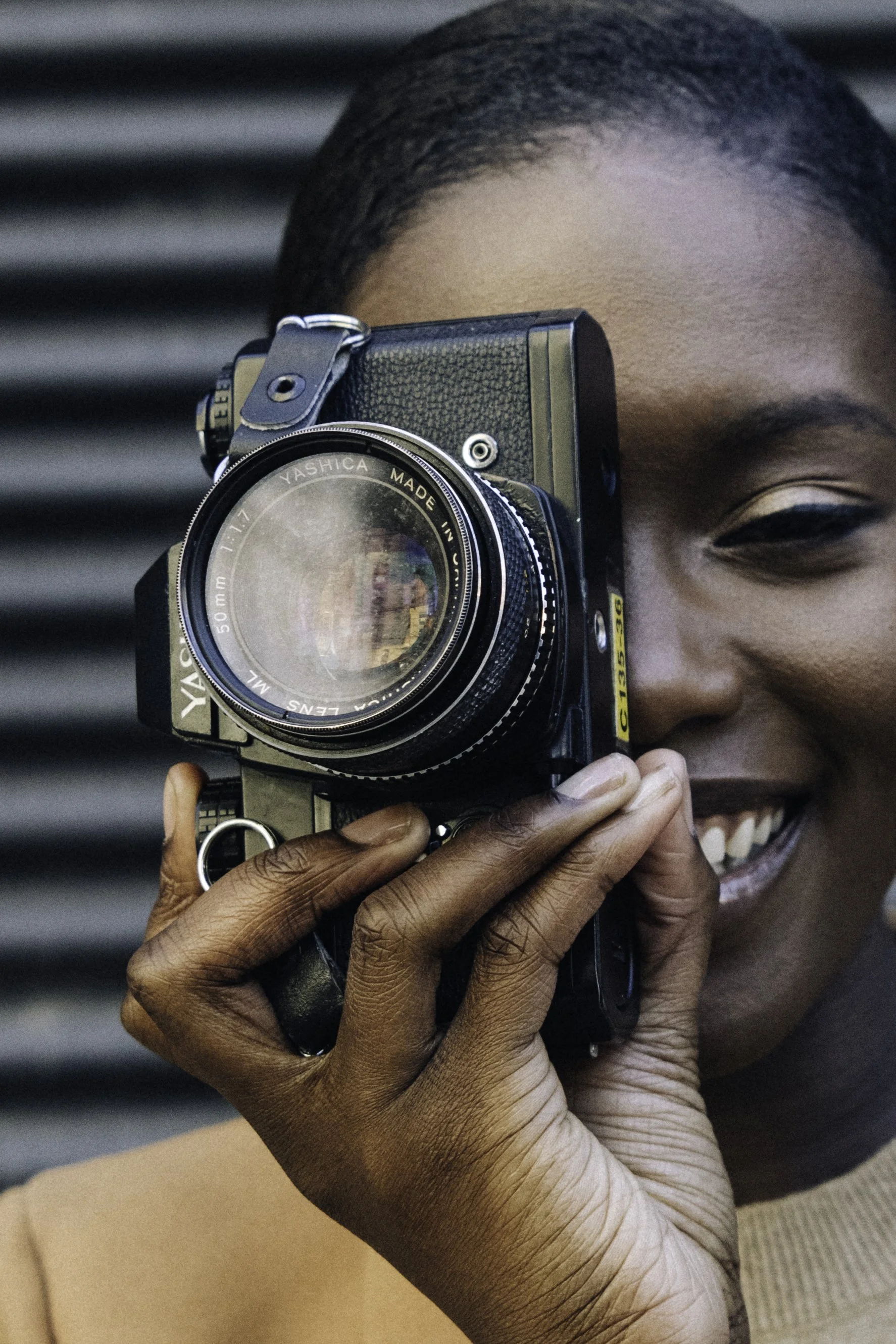 A woman holding a vintage film camera up to her face, smiling, with a dark striped background.