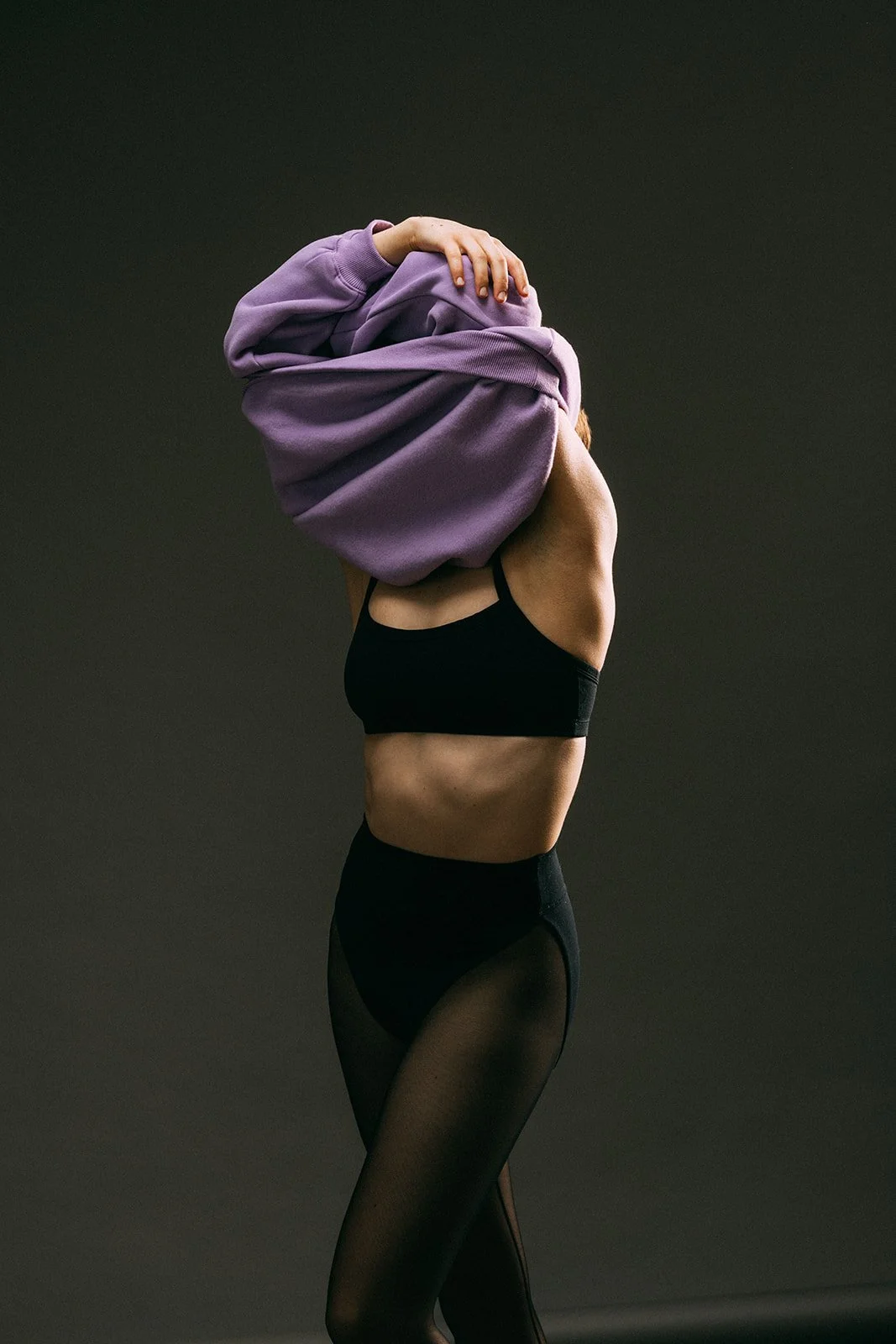 A woman wearing black sports bra and high-waisted black leggings, holding a purple hoodie over her head against a dark background. Fitness, sports, commercial photography, Chicago based photographer.