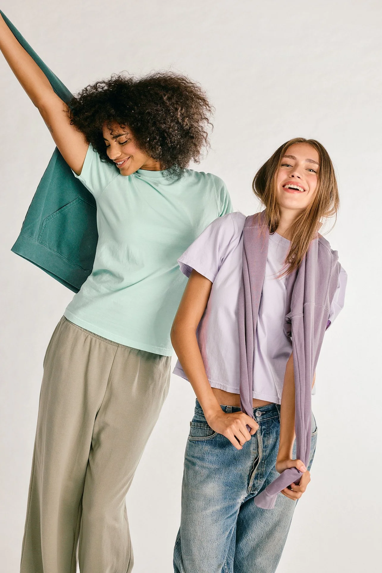 Two women smiling and posing casually against a neutral background, dressed in relaxed, casual clothing.
