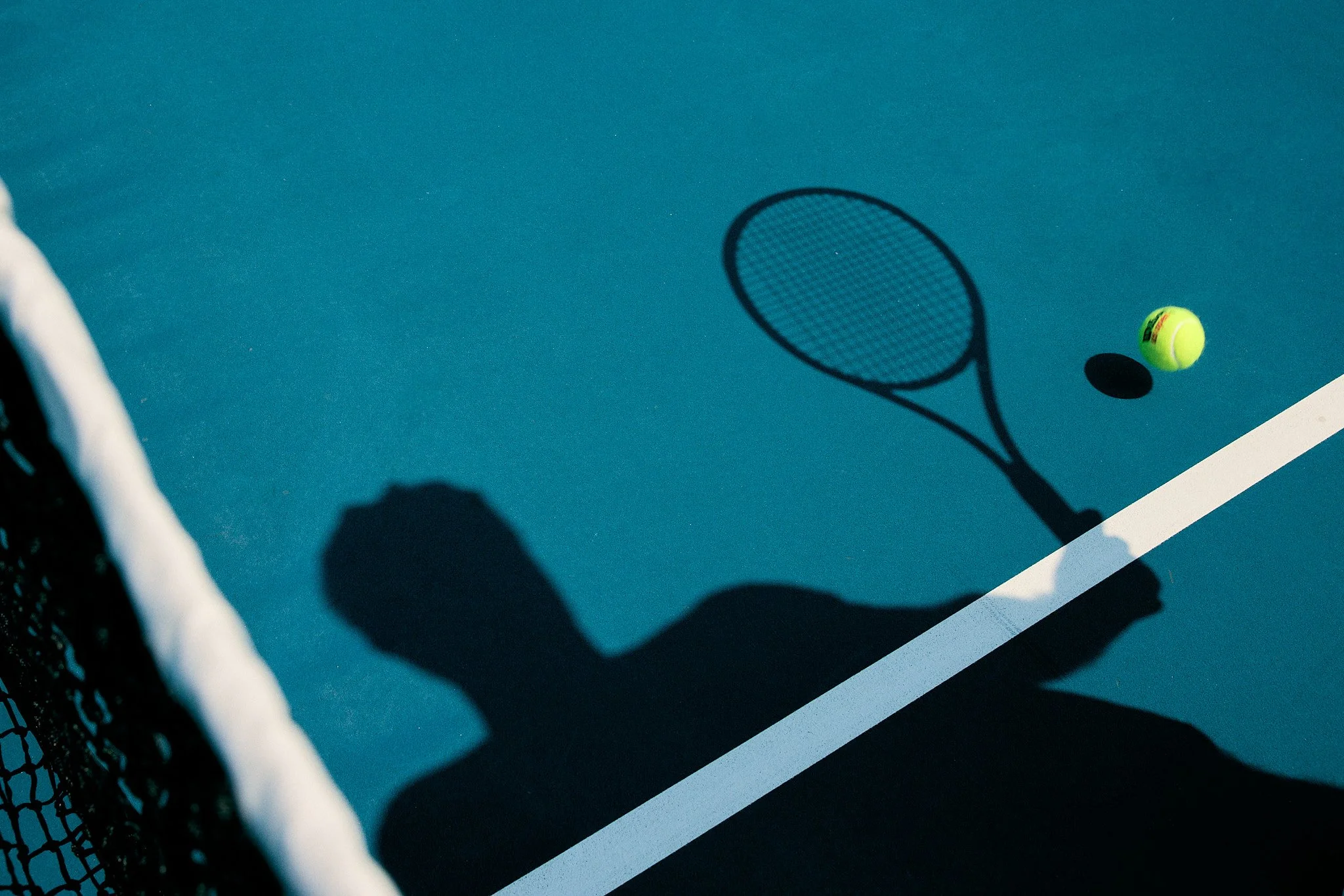 Silhouette of a tennis player with a racket on a blue tennis court, with a yellow tennis ball casting a shadow.