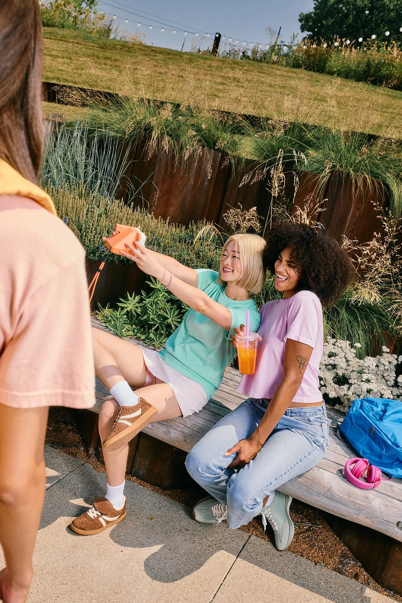 Two women sitting on a wooden bench outdoors, taking a selfie. One woman has blonde hair, wearing a mint green shirt and white shorts; the other woman has curly black hair, wearing a lavender shirt and jeans, holding a pink drink. Another person stan
