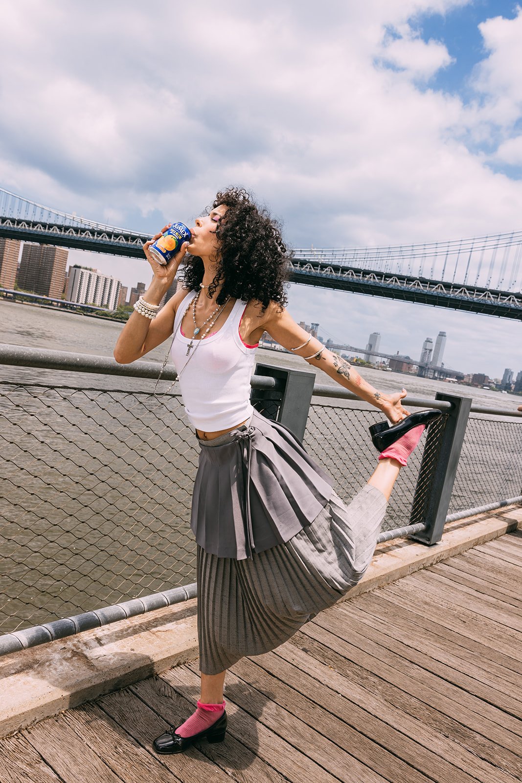 A woman with curly hair stretching her leg on a wooden deck by the water, with a bridge and city skyline in the background, holding a can of soda and striking a pose.