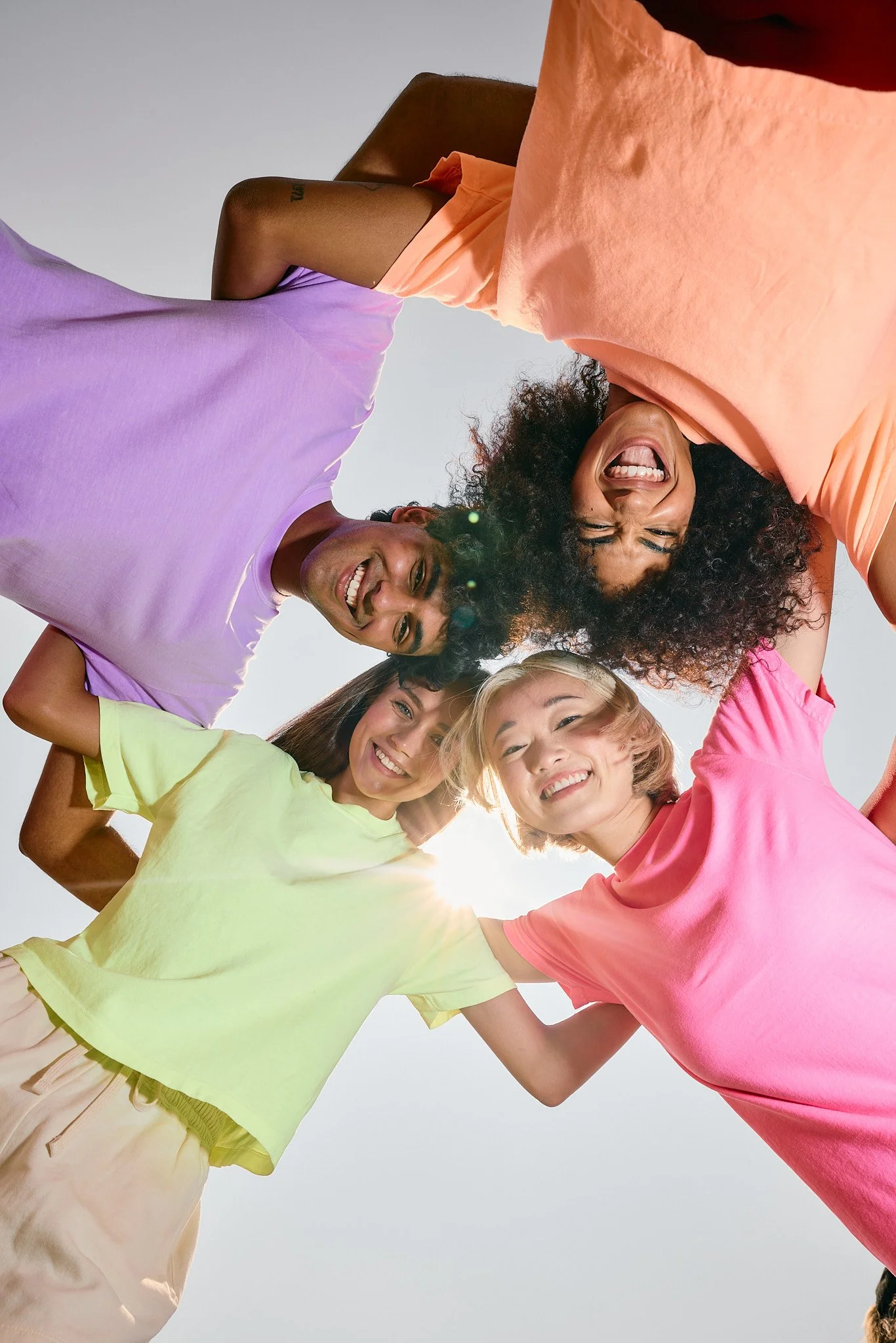 Group of five diverse people standing in a circle with heads together, smiling and enjoying outdoor sunlight.