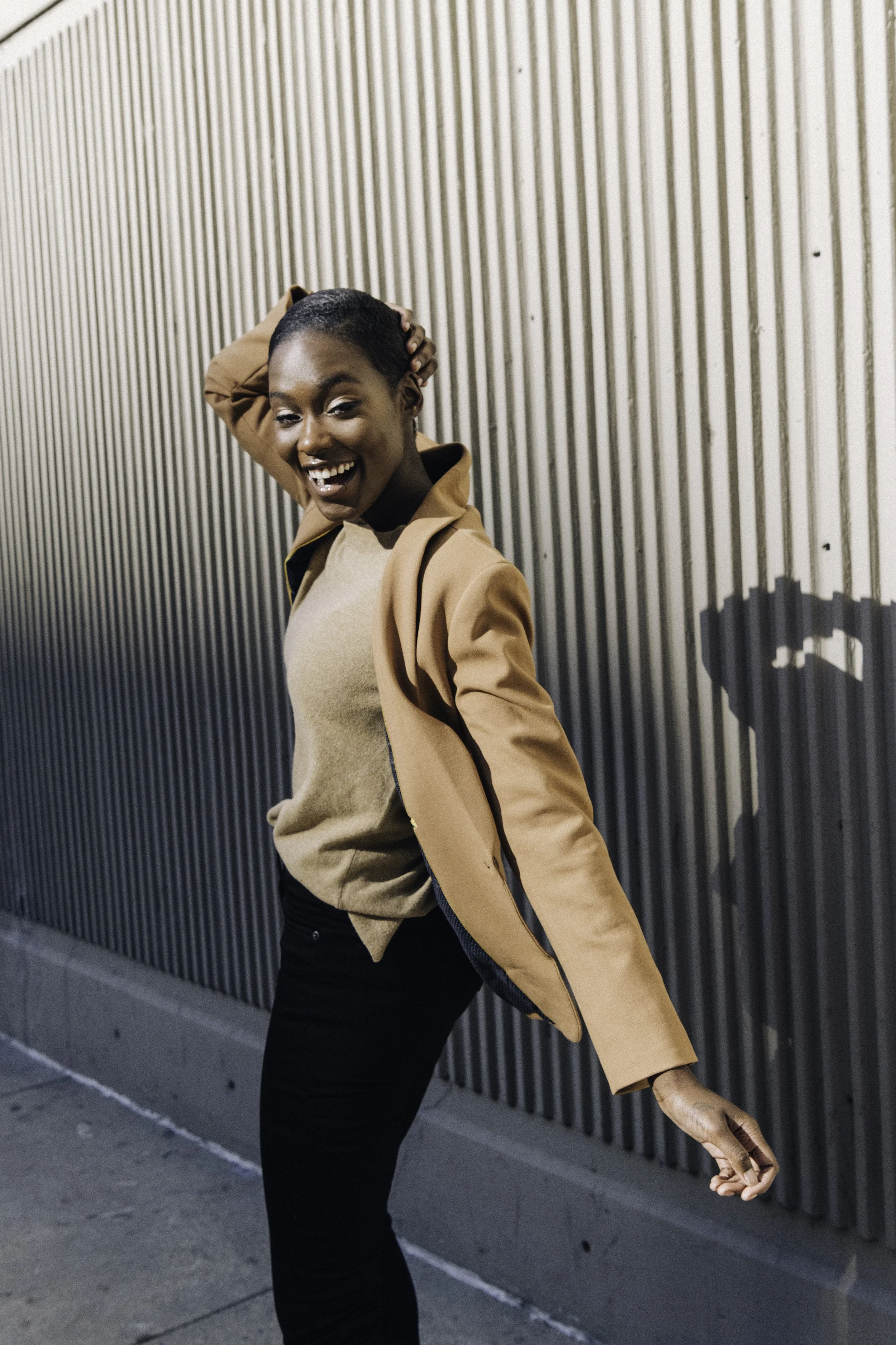 A woman in a tan jacket and black pants smiling, standing next to a corrugated metal wall, casting a shadow of a heart shape with her hand.