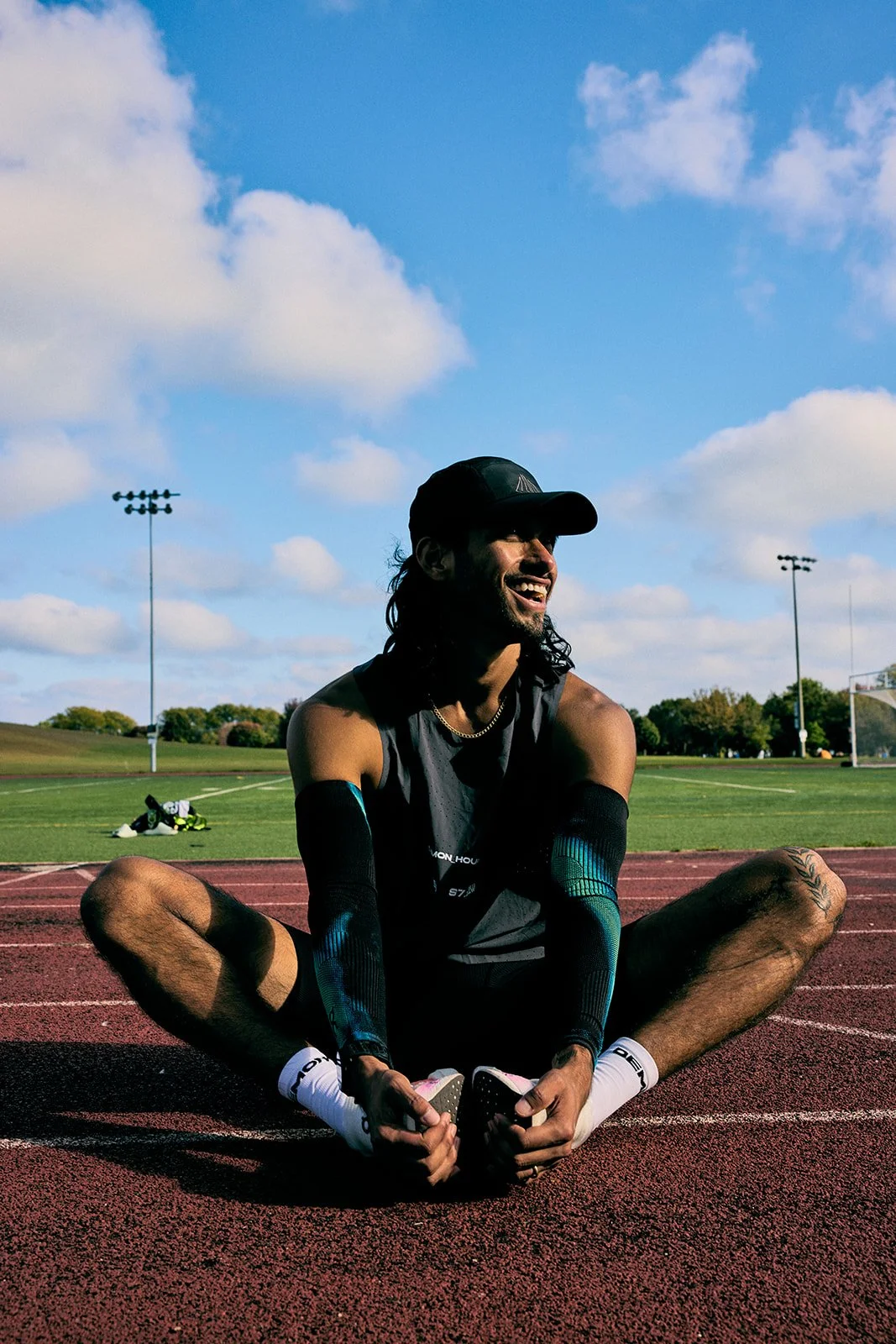 A man sitting cross-legged on a running track at an outdoor sports field, smiling and holding his shoes. The sky is partly cloudy, and there are stadium lights and a soccer goal in the background.