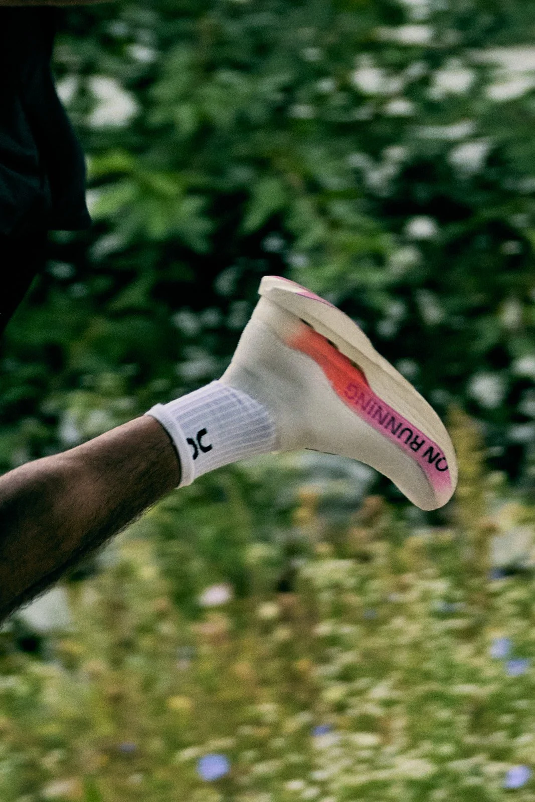 Person holding a white running shoe with pink accents, wearing a white sock with black text, in a green outdoor setting.