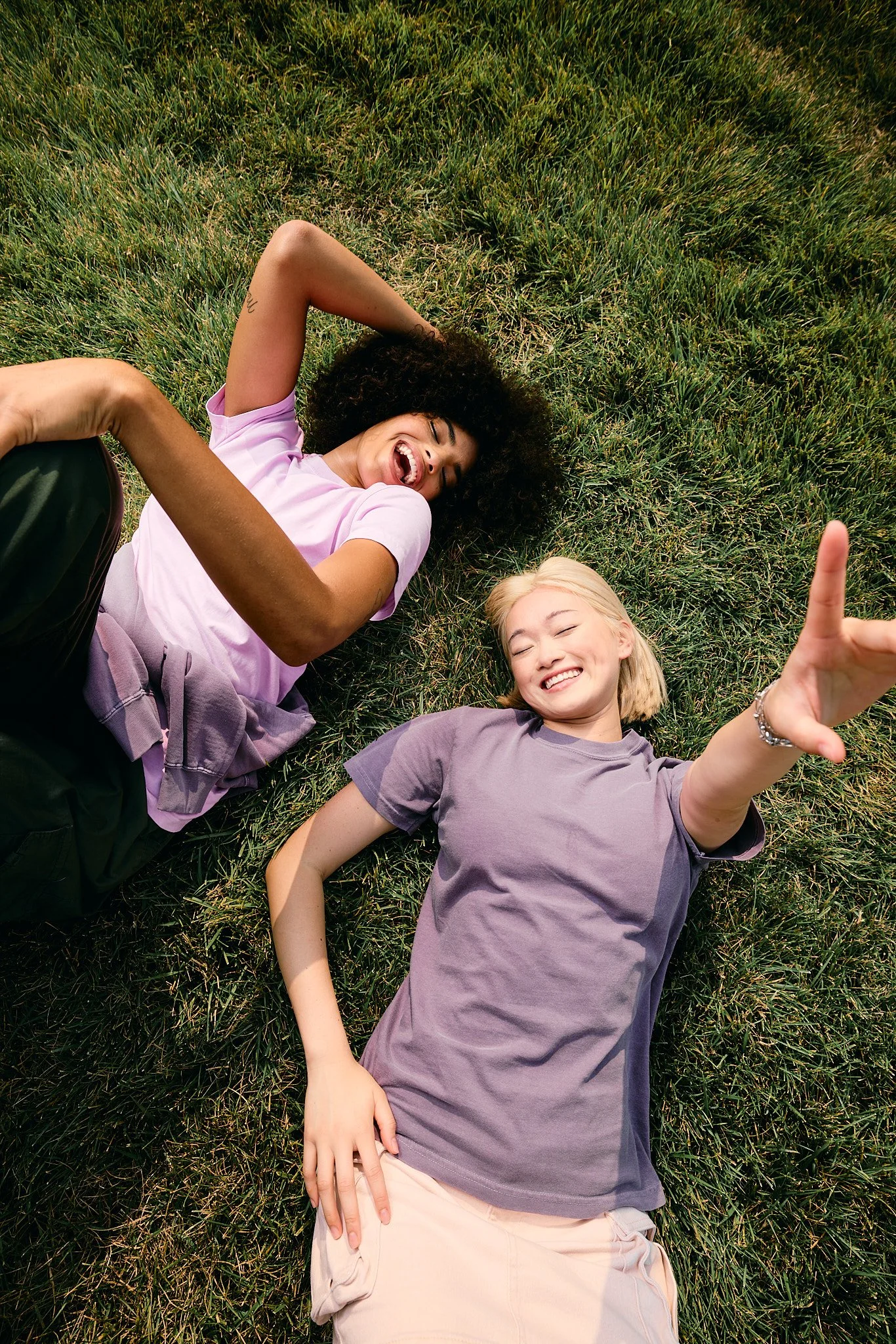 Two young women lying on grass, smiling and laughing, one with curly dark hair wearing a pink shirt, the other with blonde hair wearing a purple shirt, one pointing at the camera.