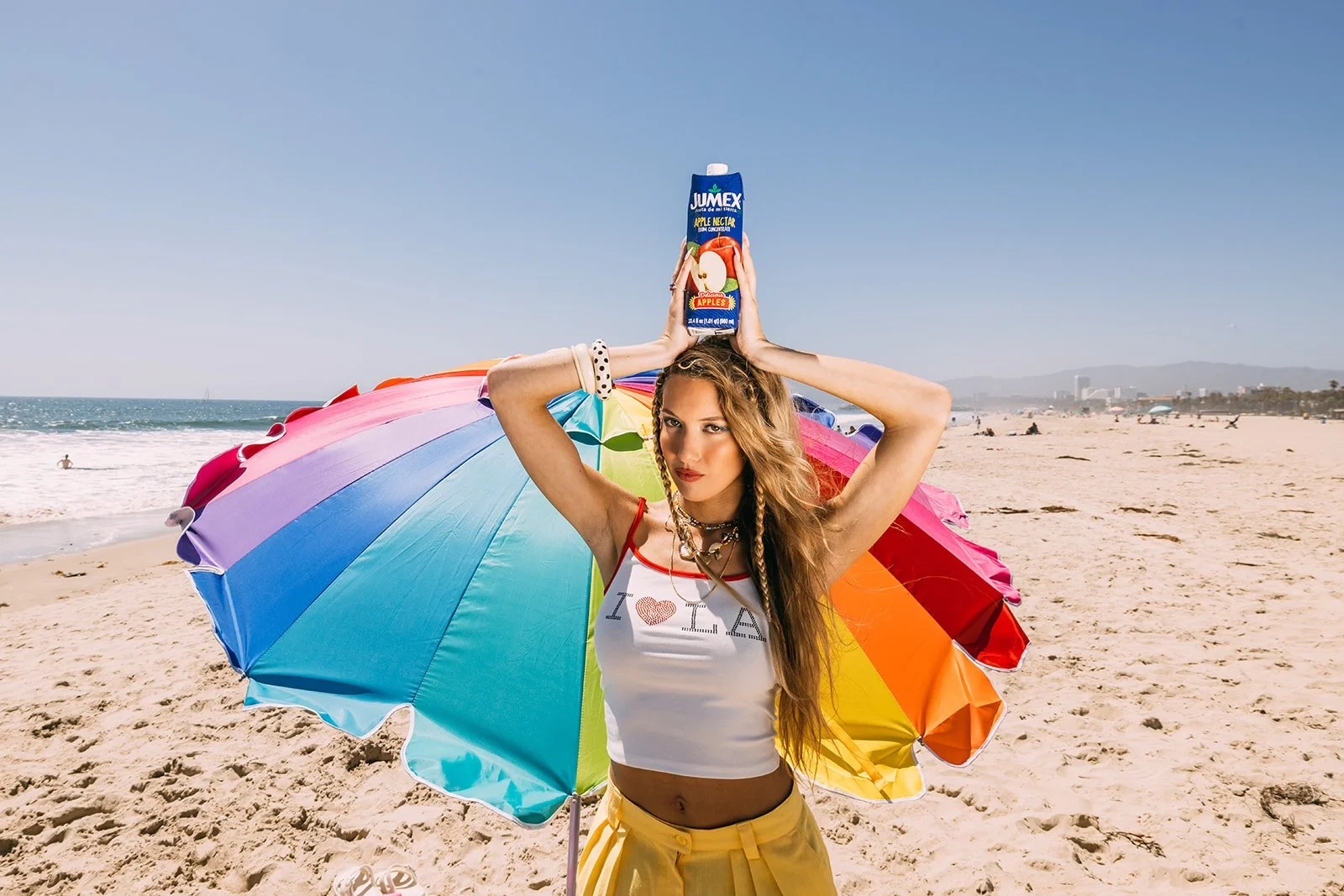 A young woman with long, wavy hair and braids standing on a sandy beach, holding a rainbow-colored umbrella behind her back, with a colorful umbrella behind her. She wears a white crop top with red trim that says 'I ❤️ LA', yellow pants, and jewelry.