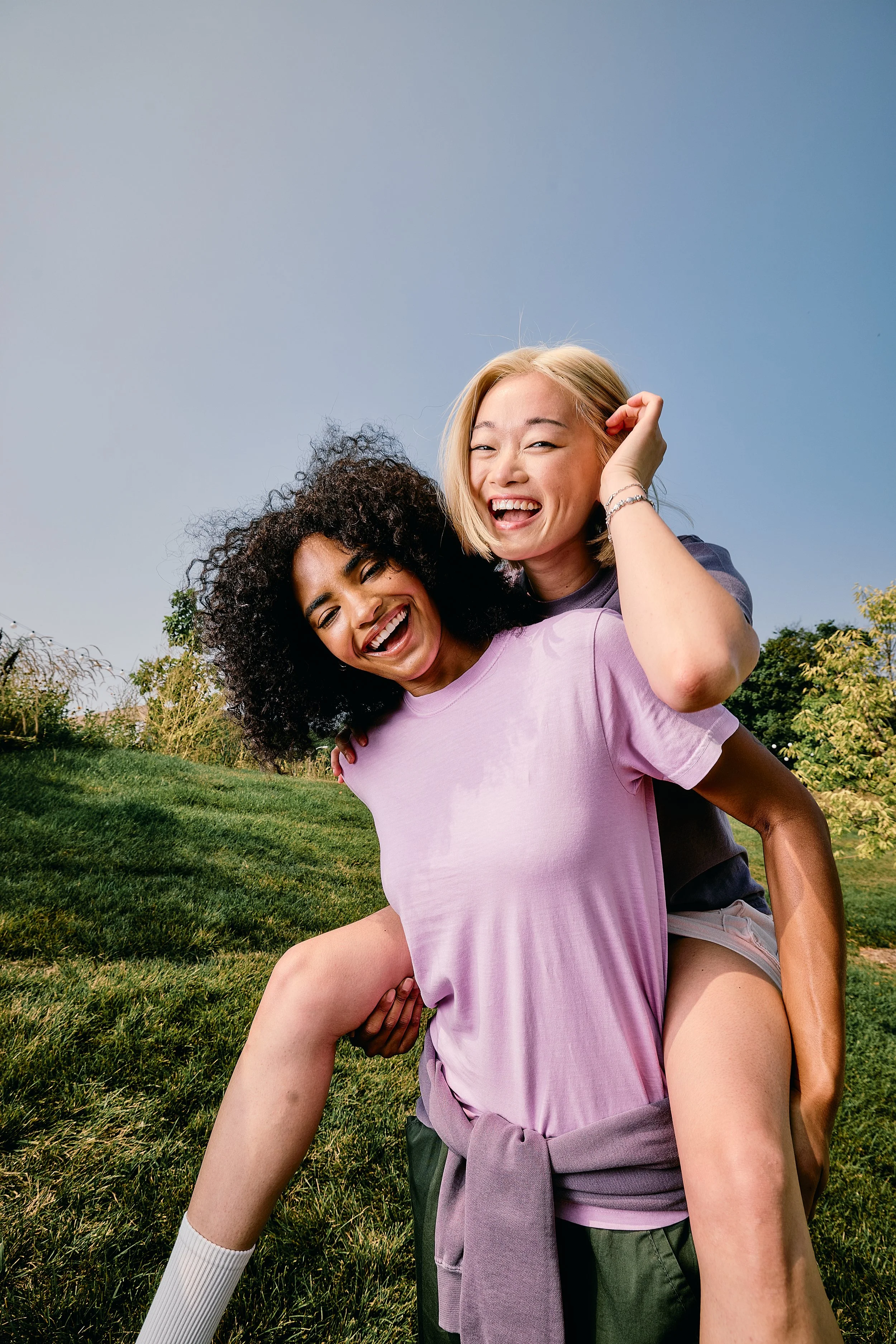 Two happy women enjoying a piggyback ride outdoors, smiling and laughing, with green grass and trees in the background under a clear blue sky.