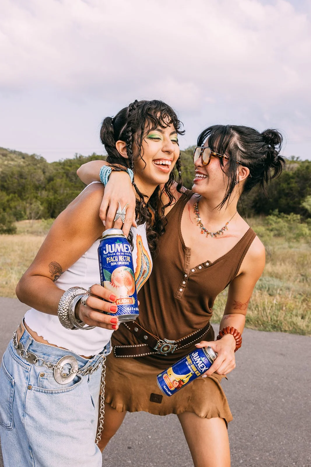 Two women smiling and laughing outdoors, holding cans of Juicy Peach Nectar drink, with grass and trees in the background.