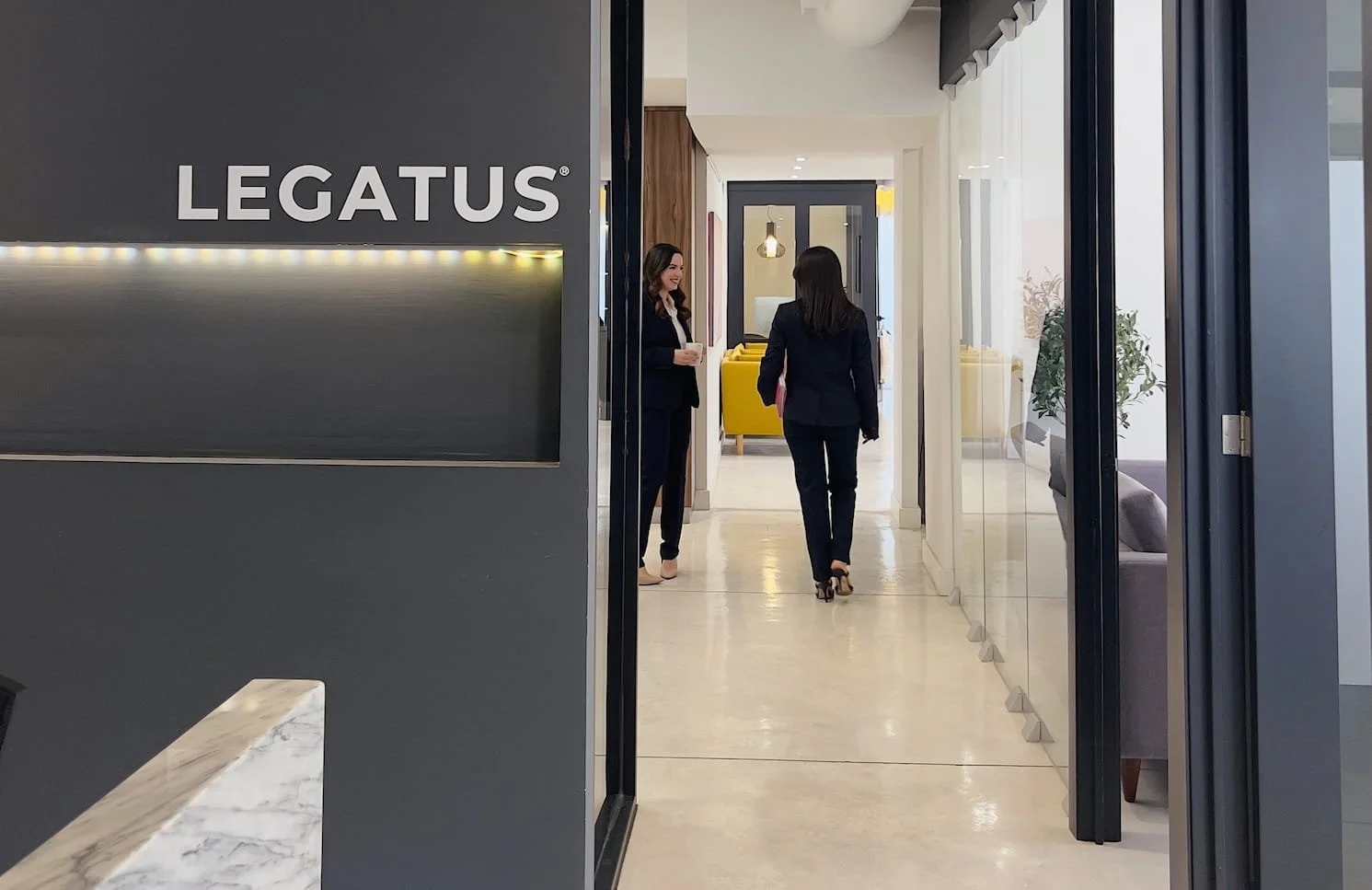 Entrada de oficina con un cartel que dice 'LEGATUS', dos mujeres vestidas con trajes formales conversando en el interior, ambiente moderno y bien iluminado.