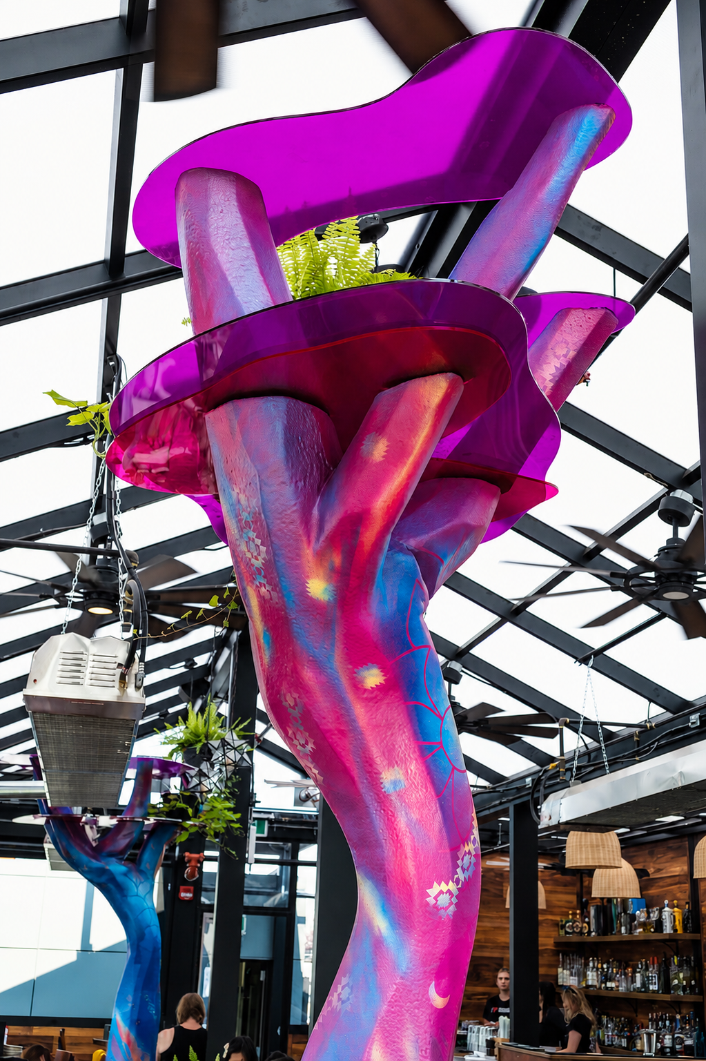 A large, colorful tree-shaped structure with purple, blue, and pink hues, serving as an interior decor element in a restaurant or cafe, with plants on the branches and a bar area with bottles in the background.
