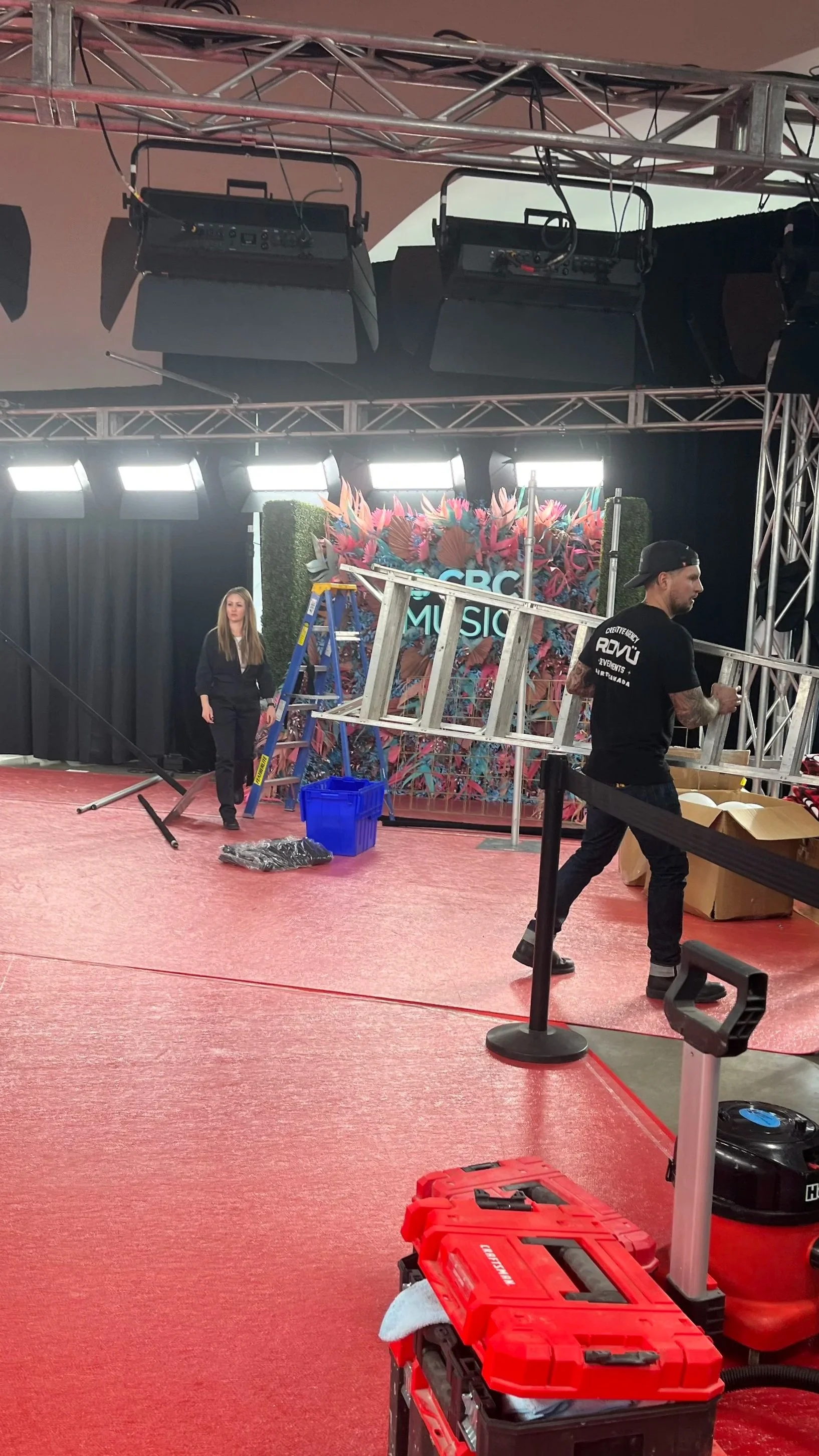 Event setup with stage, metal scaffolding, banners, and lighting equipment. Two workers installing or preparing the stage, with tools and storage boxes in a large indoor venue.