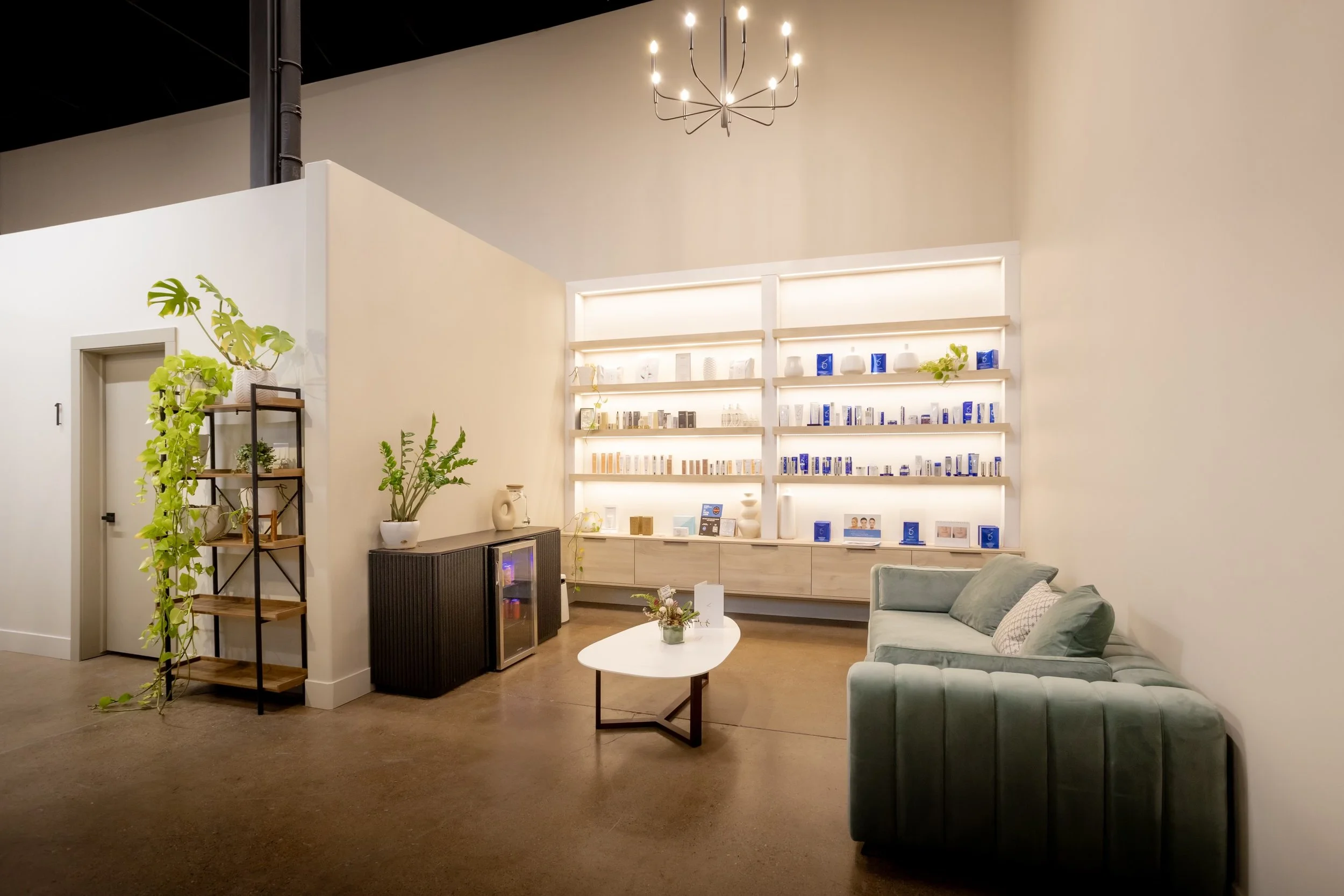 A modern waiting or lounge area with a beige couch, a white coffee table with a small plant, a gray patterned armchair, and a floating wall shelf displaying decor and skincare products. There are green plants in pots, a black mini fridge, and a decorative black metal and wood shelving unit. The upper ceiling features a black modern chandelier with multiple exposed bulbs.