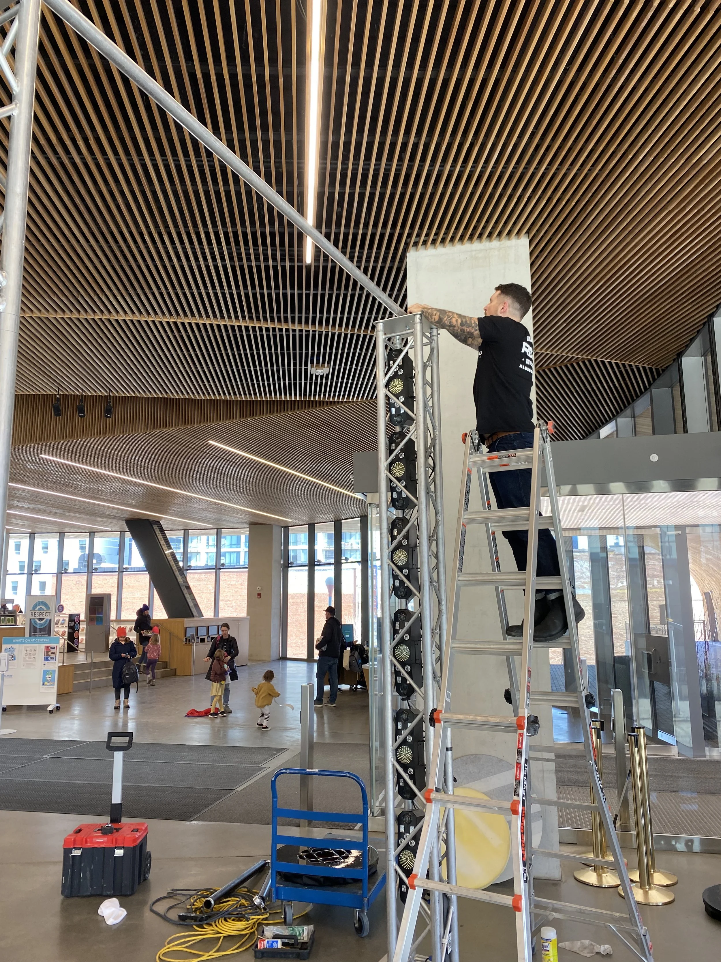 A man is standing on a ladder working on a ceiling in a large, modern indoor space, likely a public building or airport, with people walking and children playing in the background.