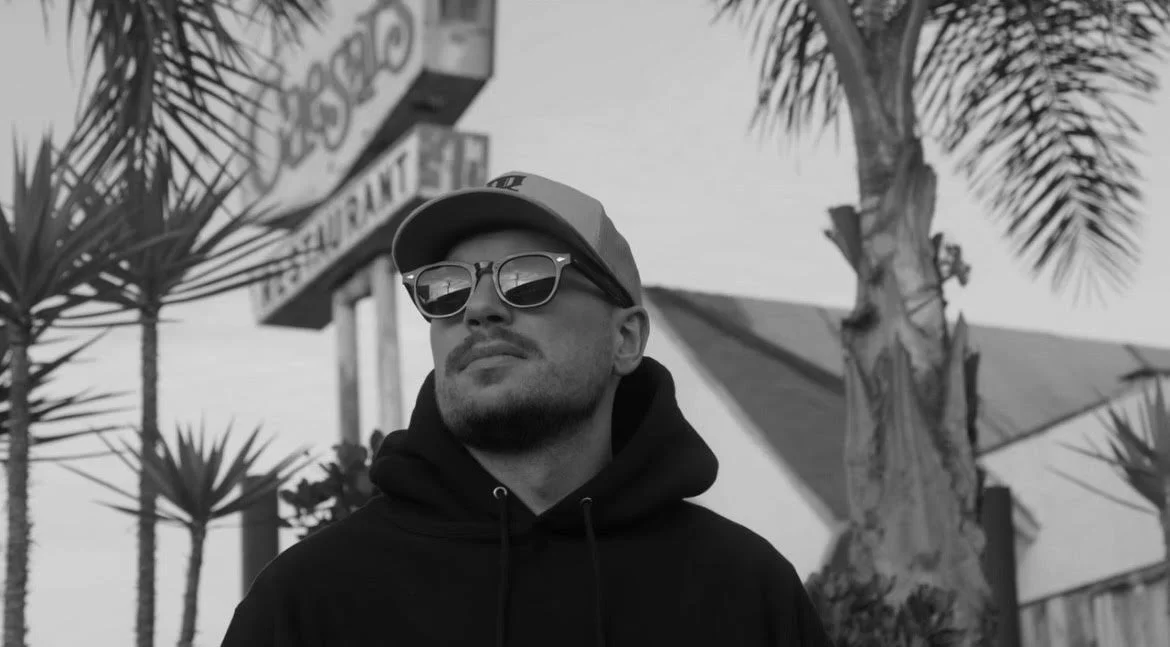 A man wearing sunglasses, a cap, and a hoodie standing outdoors, with palm trees and a building with a sign in the background.