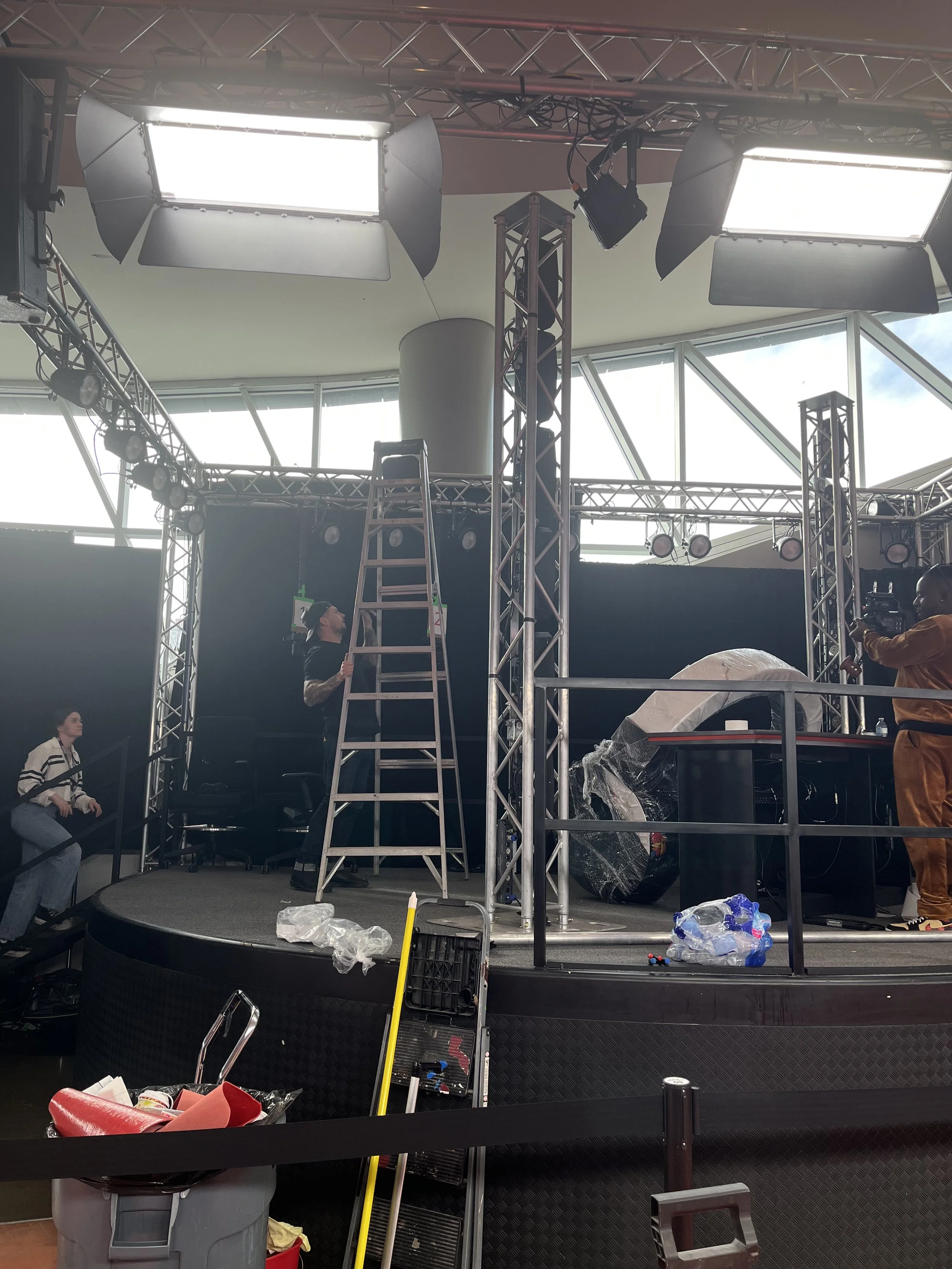 Stage being set up with lighting equipment, ladders, and crew members working, in an indoor venue with large windows.