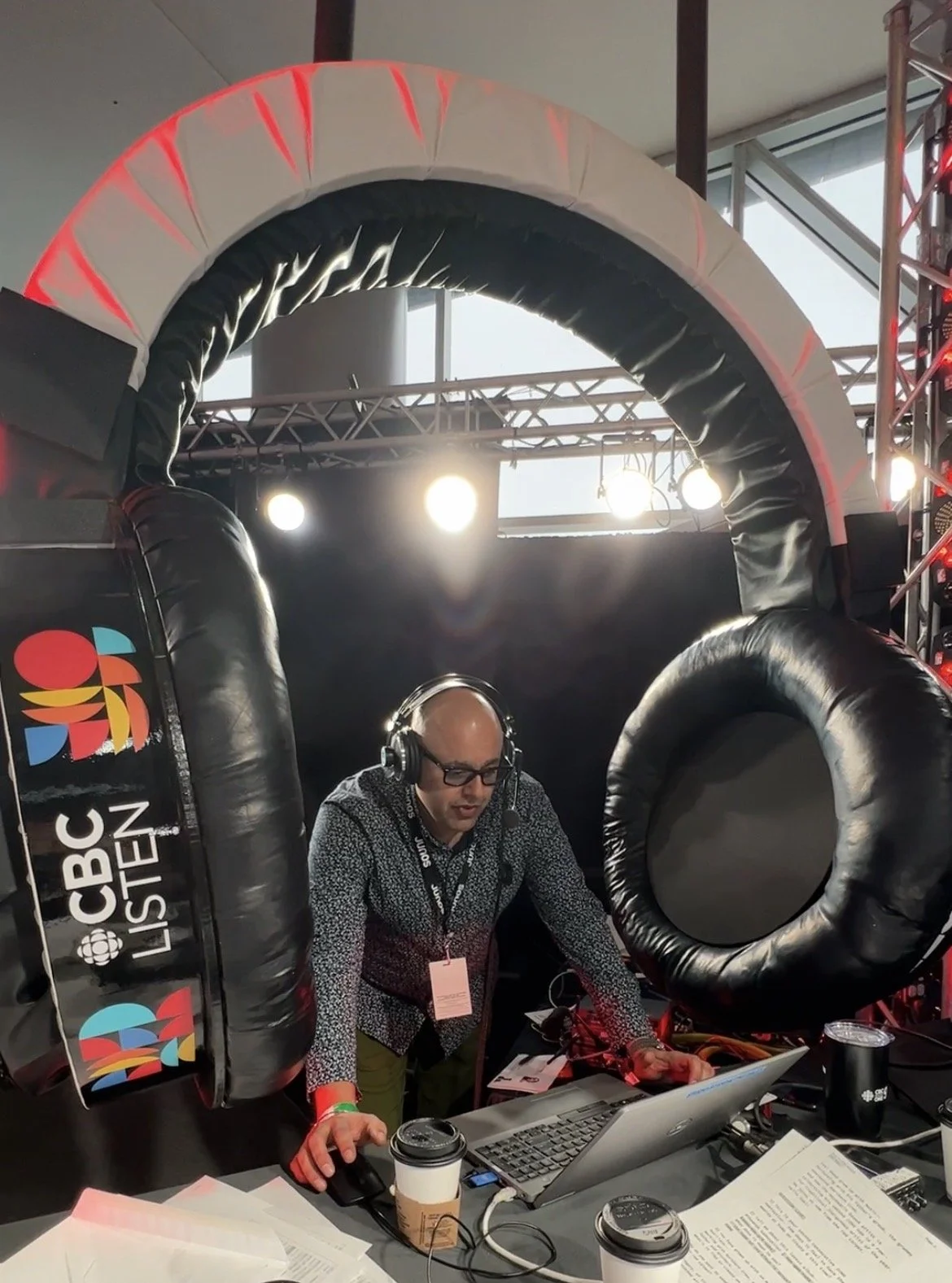 A man wearing glasses and a lanyard with a press badge, operating audio and computer equipment at a broadcast station for CBC News, with a backdrop of stage lights and large inflatable branded archway.