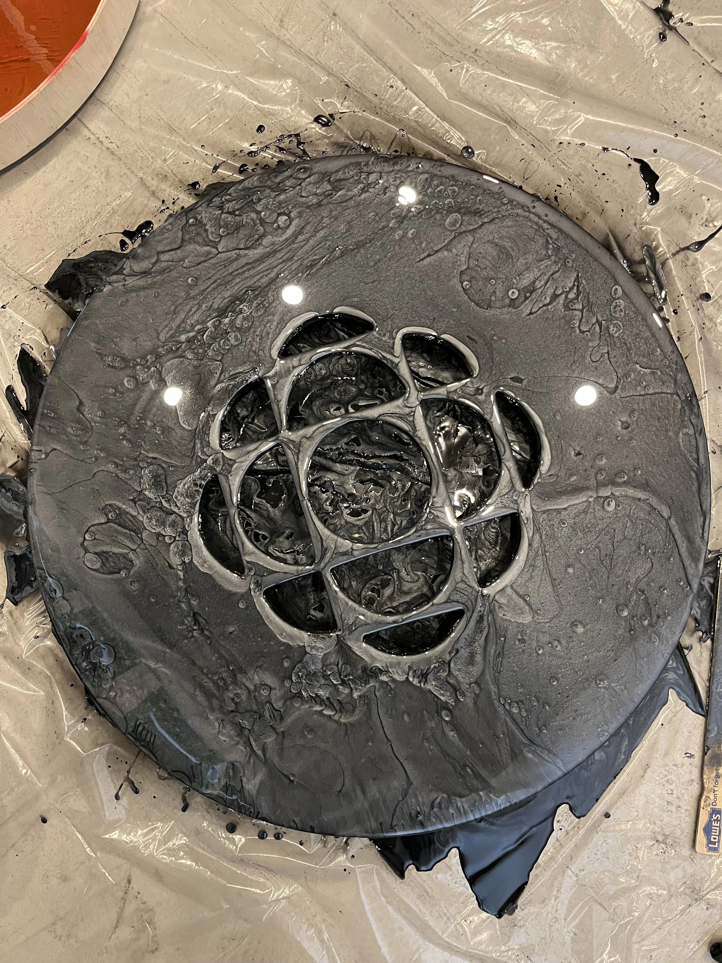 A circular object with a spider-like pattern in the center, covered with black liquid paint, on a plastic sheet on the floor