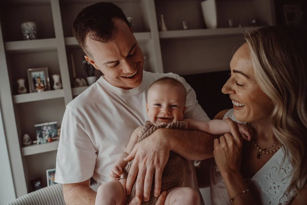 A happy family consisting of a man, a woman, and a smiling baby in a cozy room with decorative shelves in the background.