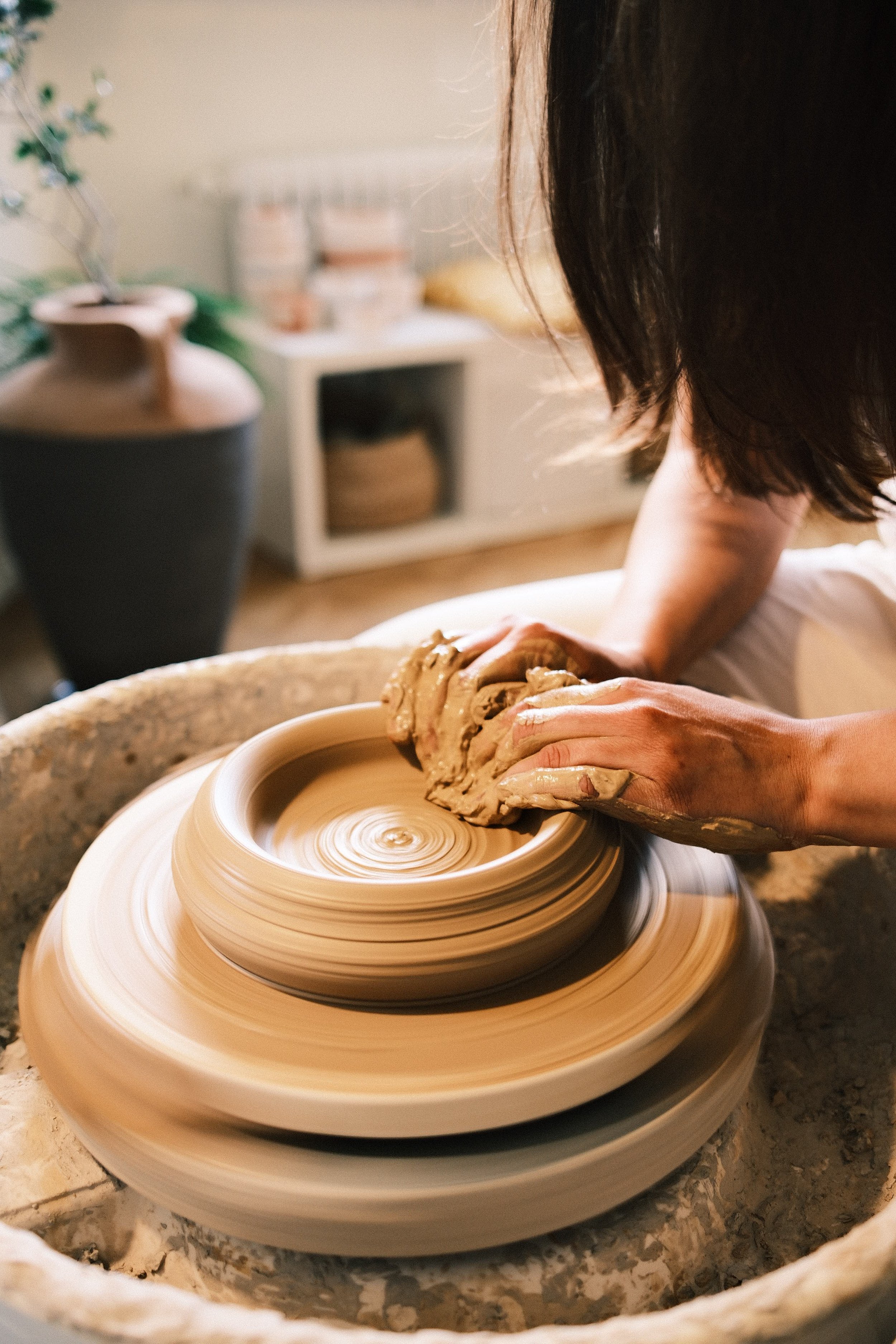 A person shapes clay on a spinning pottery wheel in a pottery studio, with shelves and a potted plant in the background.