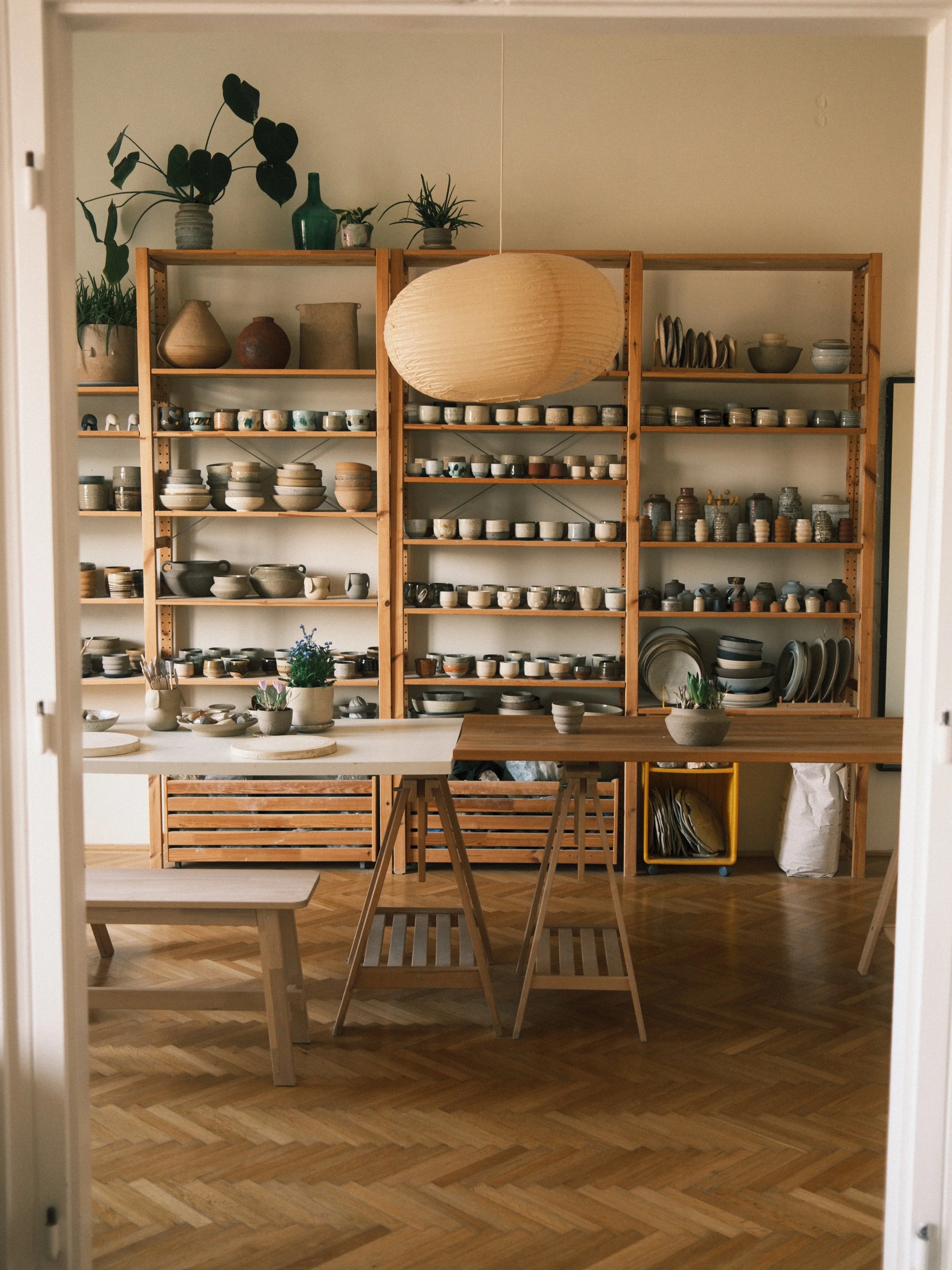A room with wooden bookshelves filled with various ceramic bowls, plates, and pots. Two small potted plants and a large paper lantern hanging from the ceiling are visible. There are wooden chairs and tables in the foreground.