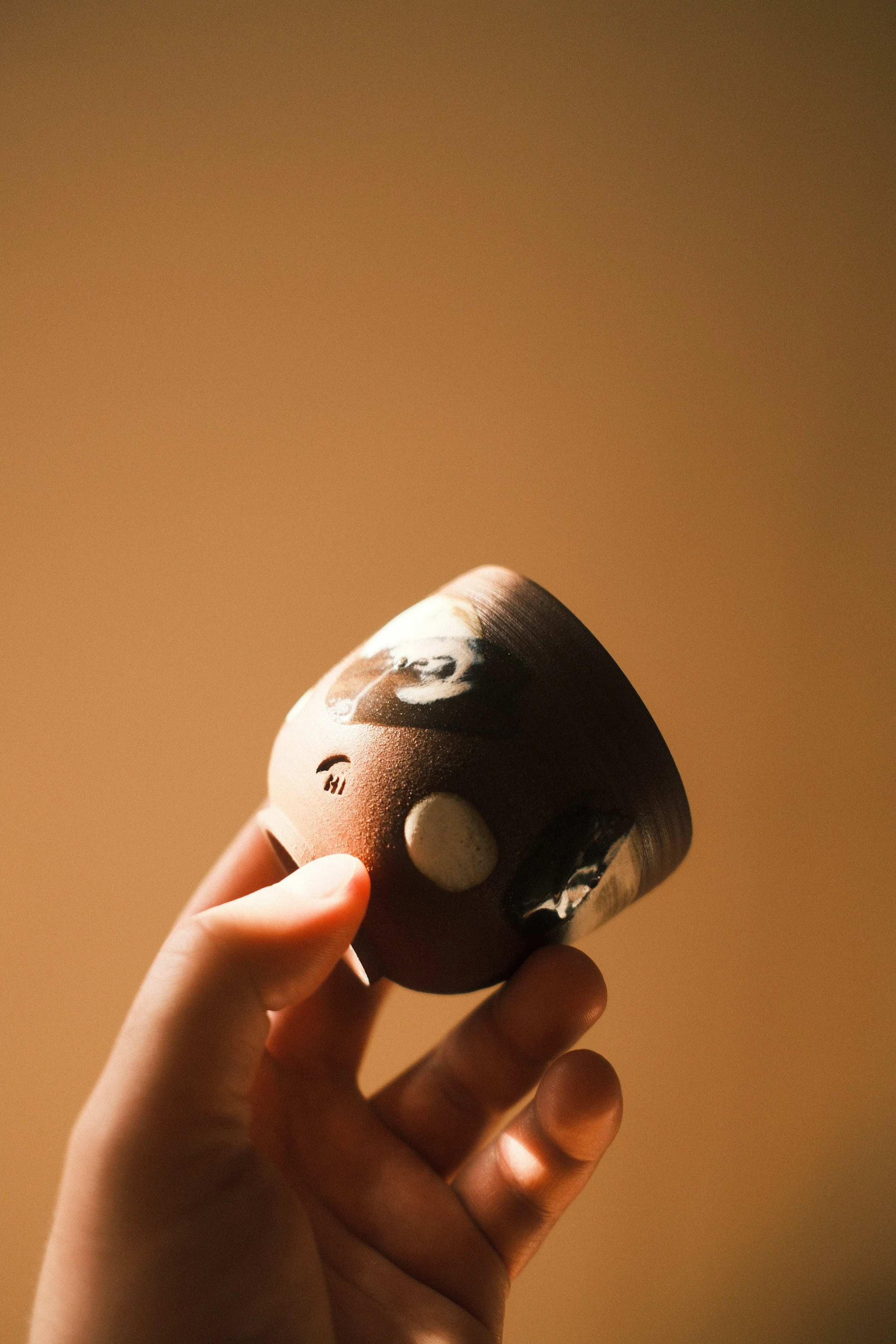 A person's hand holding a small animal-shaped ceramic figurine with a brown and black color pattern and white spots, against a plain beige background.