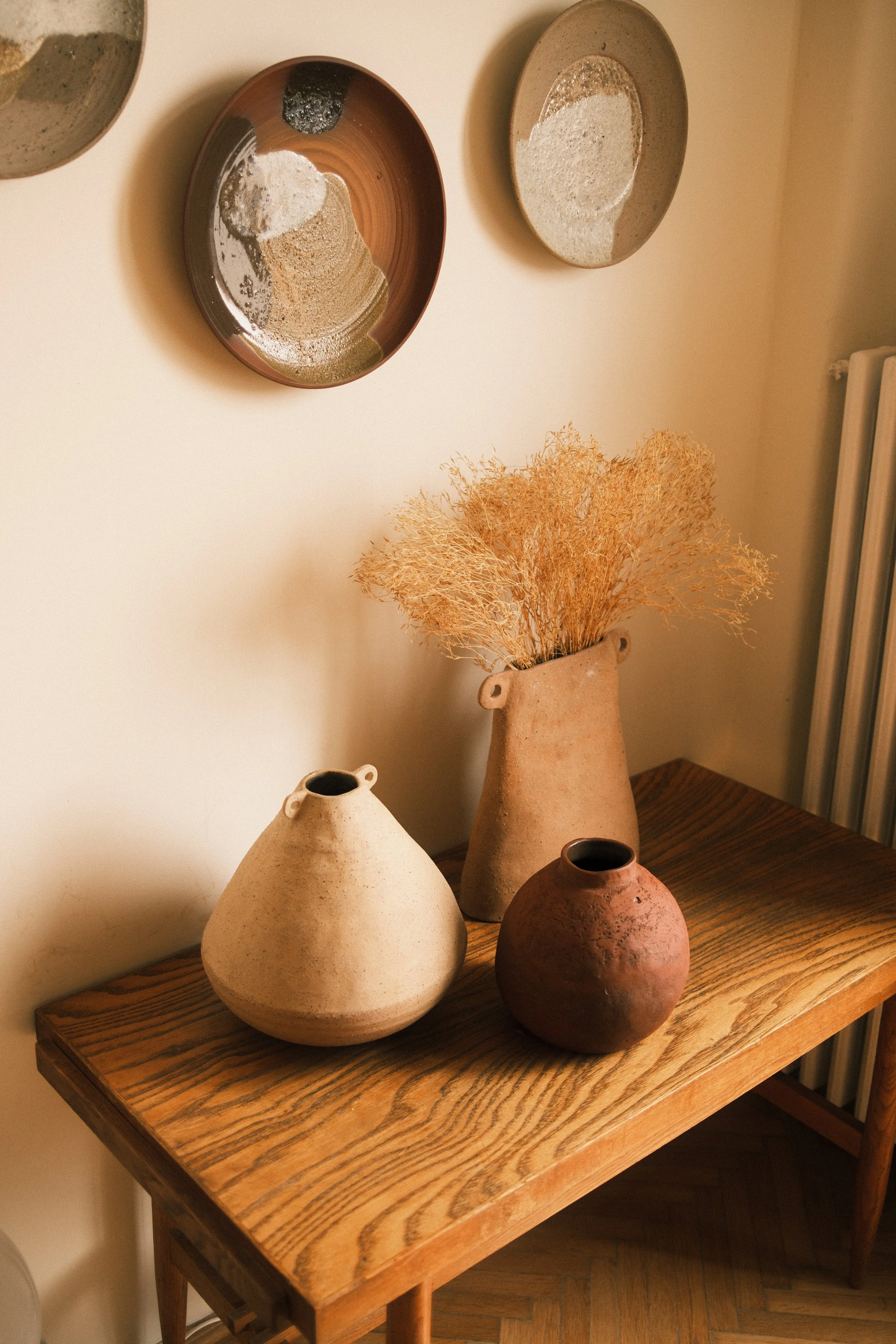 A wooden table holding five ceramic vases of various shapes and sizes, with dried decorative grass in a tall vase, against a plain wall with three decorative plates hanging on it.