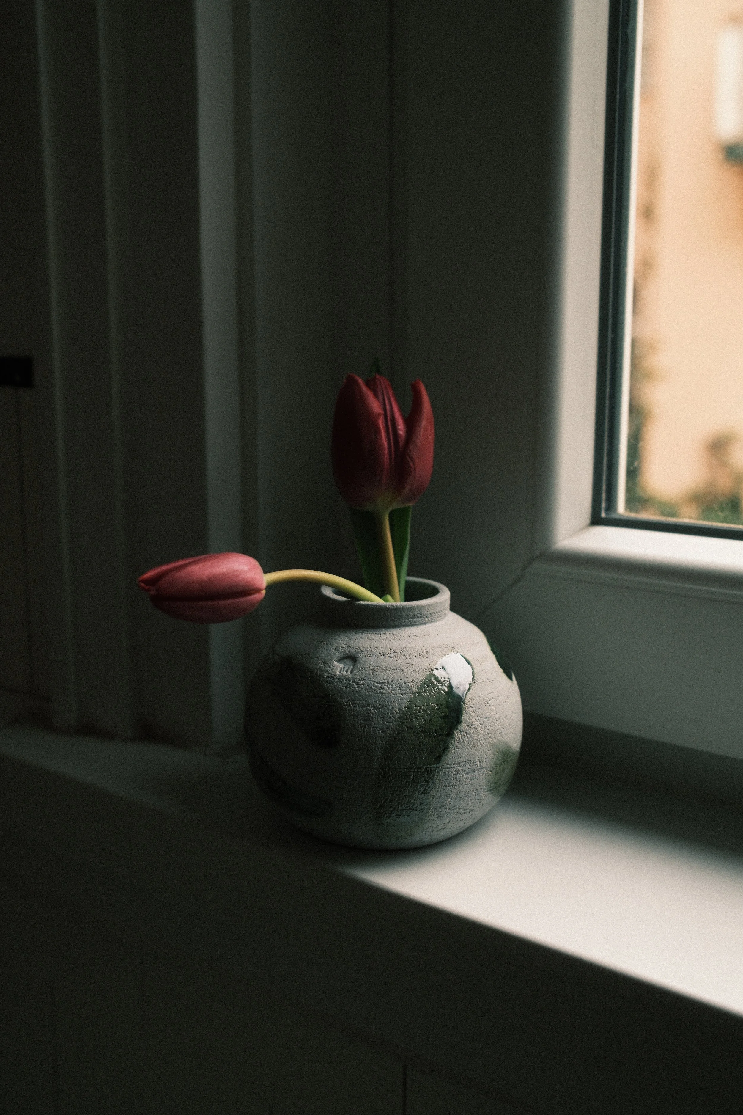 A white ceramic vase with a rough texture, holding three tulips: two pink and one dark red, placed on a windowsill with natural light and a window frame in the background.