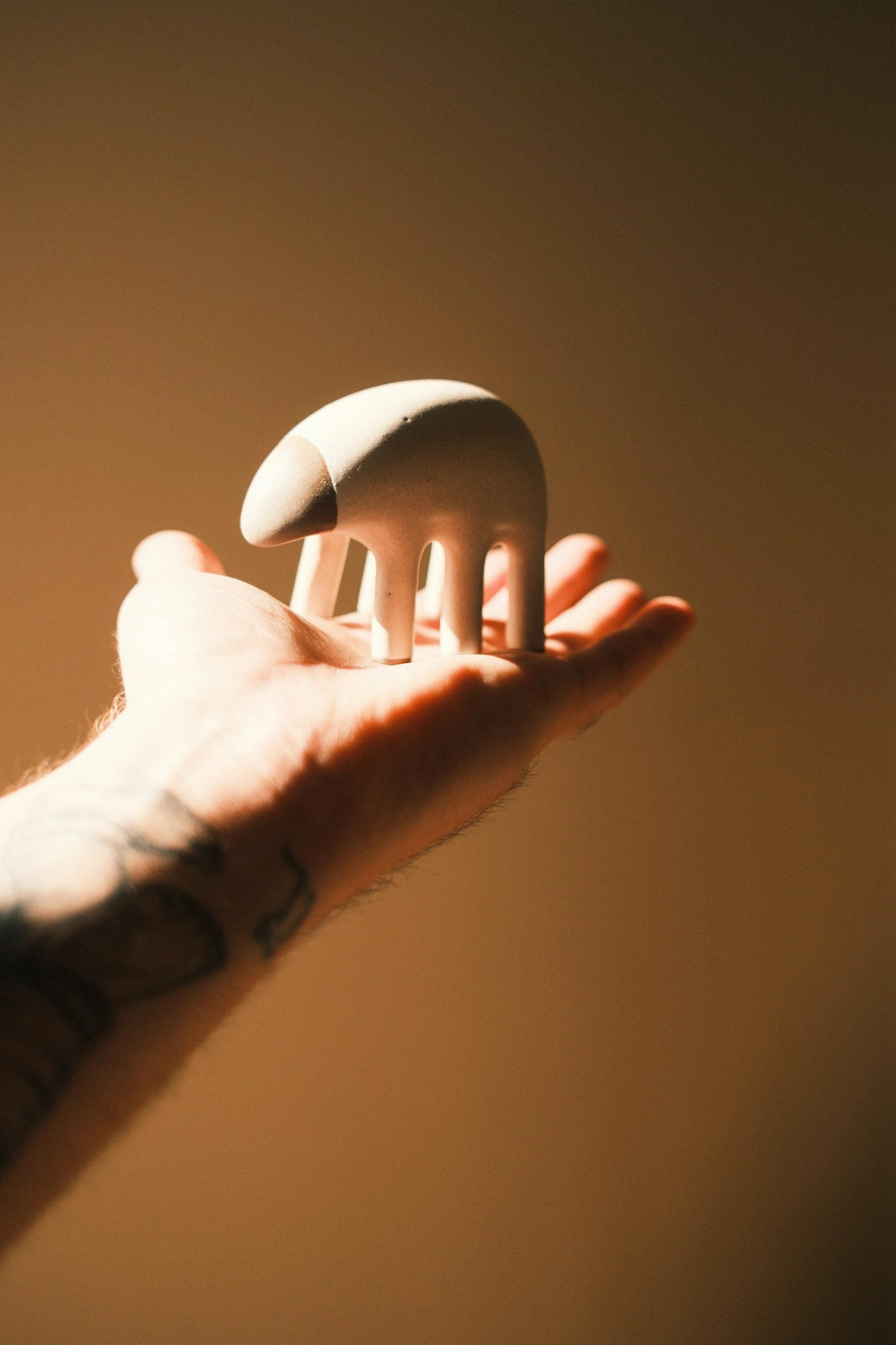 A hand holding a small, rounded, pale-colored sculpture with four elongated legs, set against a plain brownish background.