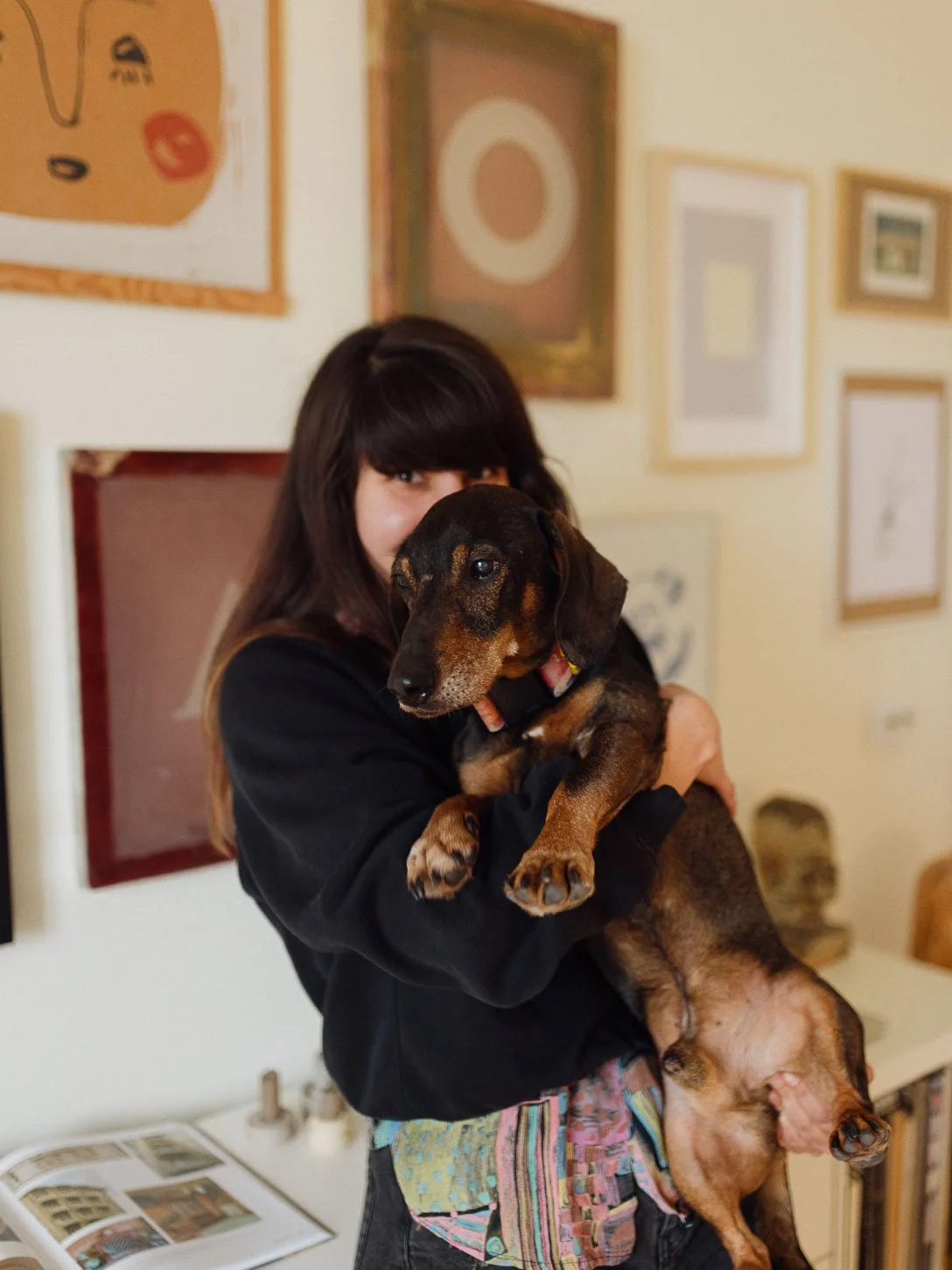 A woman holding a brown and black Dachshund puppy in an art gallery with various framed artworks on the wall behind her.