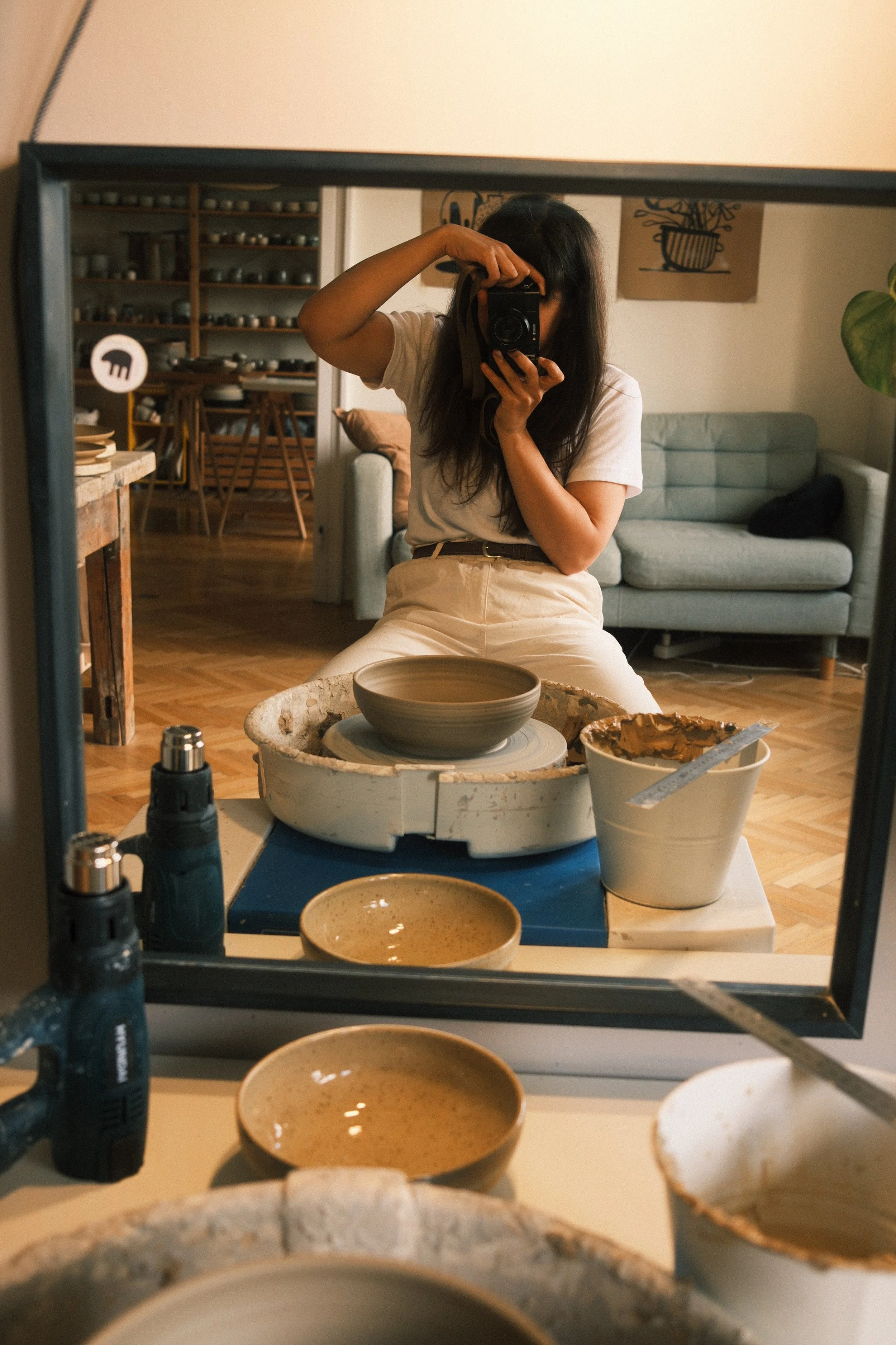 A woman with long dark hair taking a photo of herself with a camera in a mirror in her living room. The room has a big gray sofa, wooden flooring, and a shelf with small containers in the background. The foreground shows pottery tools, bowls, and a pottery wheel on a table, indicating she is in a pottery studio or workshop.