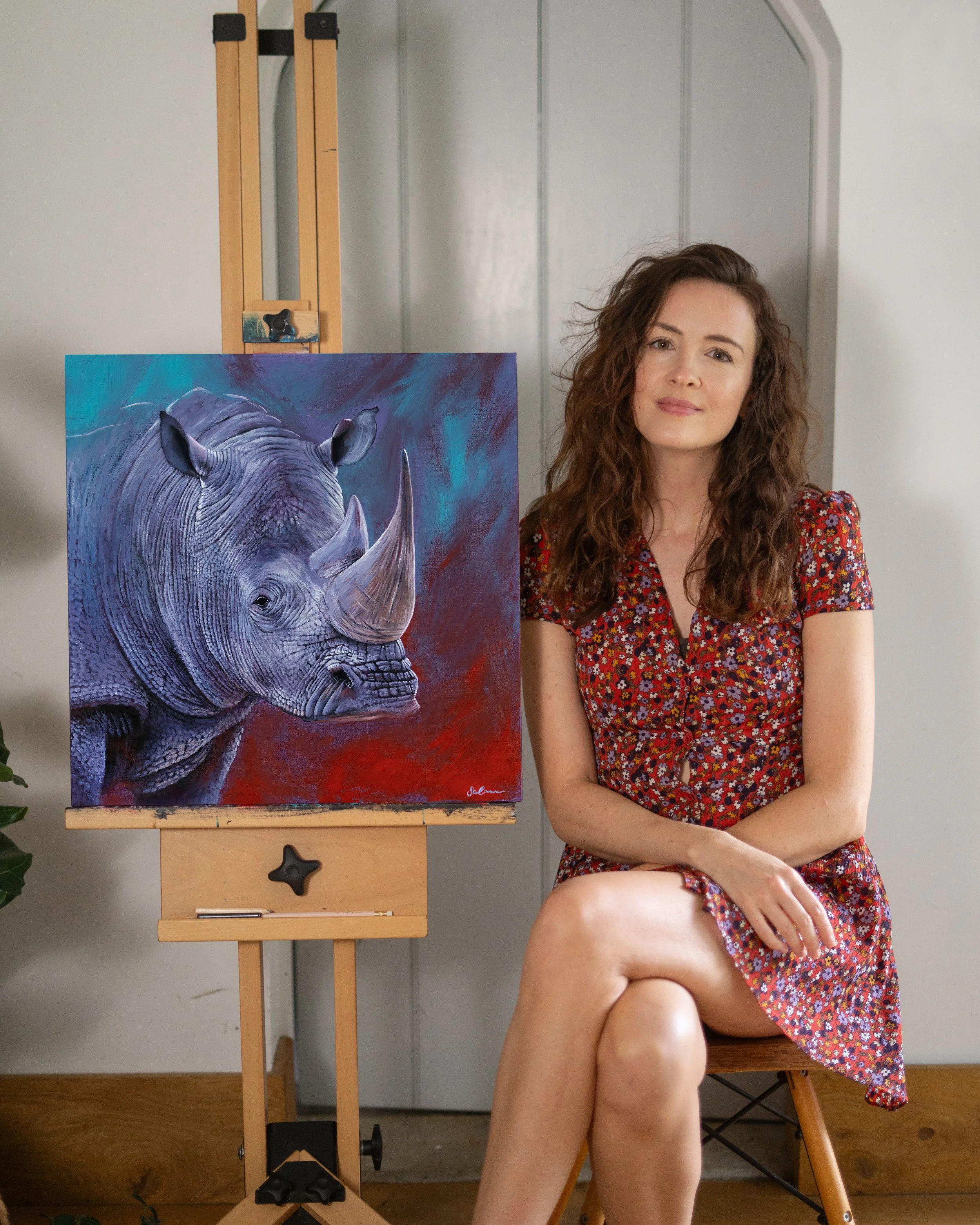 Contemporary artist, Sophie Green, sits next to a painting of a colourful rhinoceros