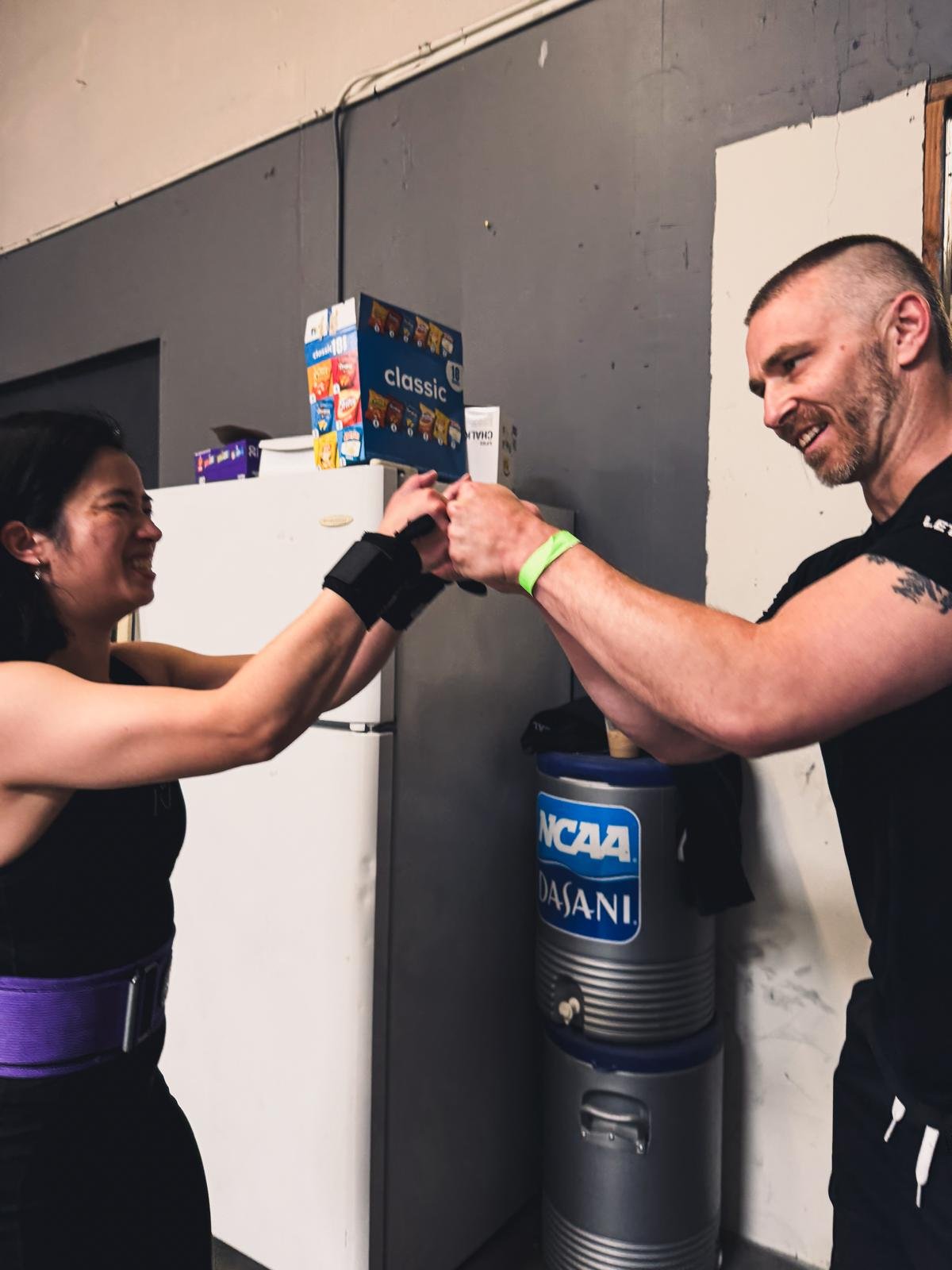 A woman and man are fist bumping in a gym, smiling, with a refrigerator and water cooler in the background.