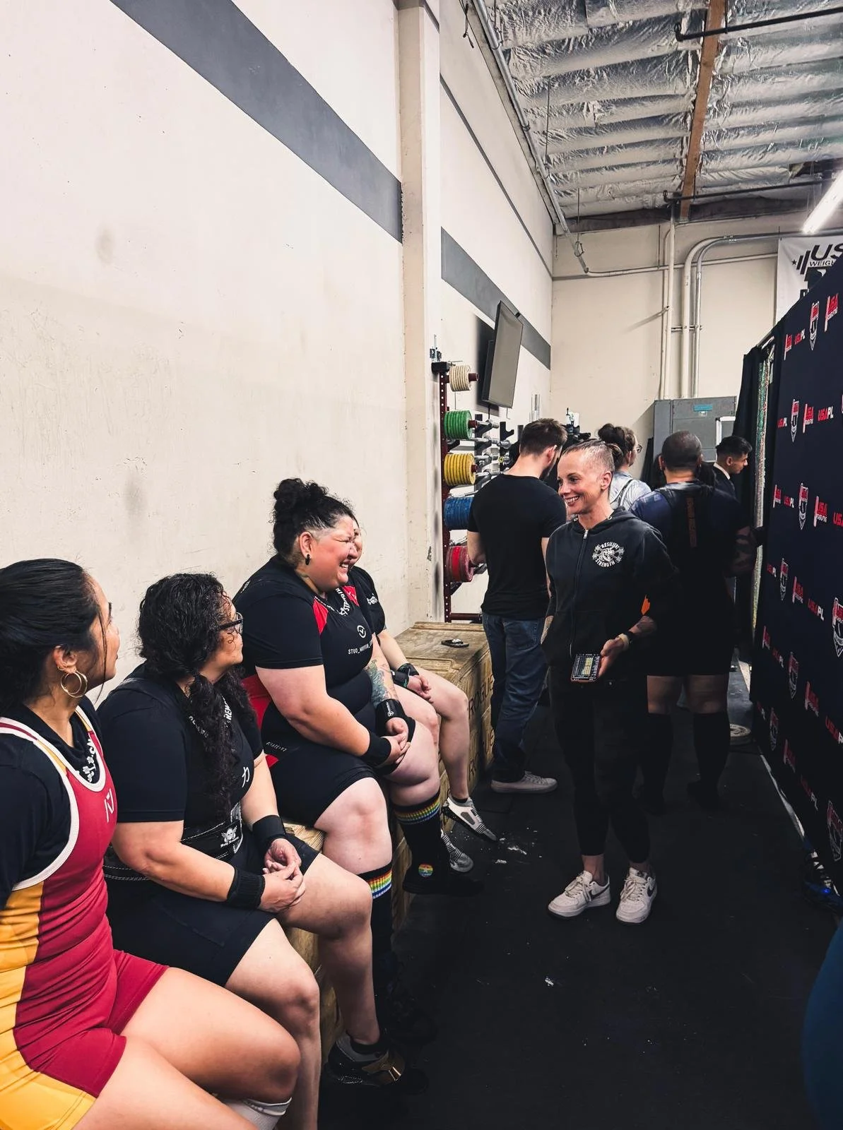 Women sitting on a bench backstage at a fitness event, smiling and talking with staff members, in what appears to be a gym or fitness venue.