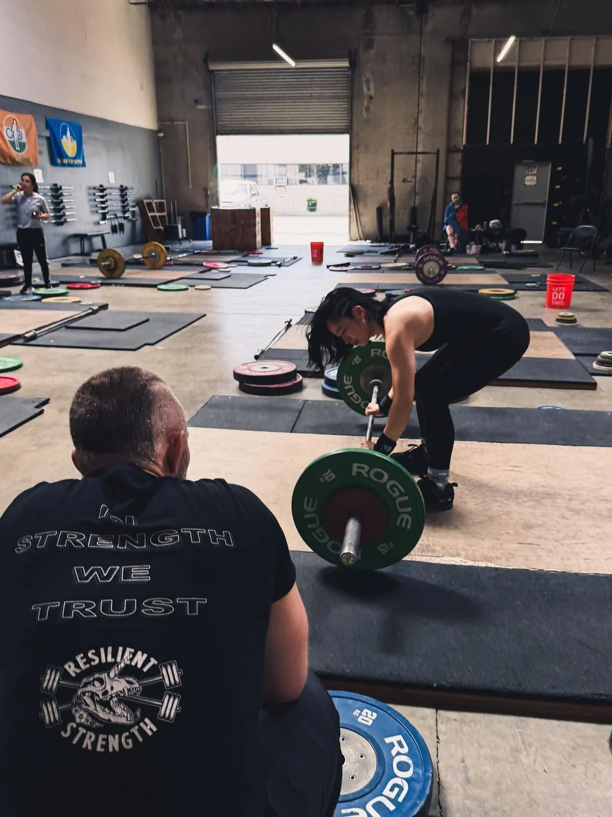 A woman lifting a barbell with green weight plates in a gym with other people working out and exercise equipment around.