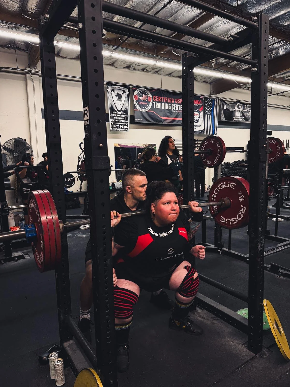 A young woman squatting in a gym, lifting a barbell with red weight plates, being assisted by a spotter. Other gym members are in the background, and banners hang from the ceiling.