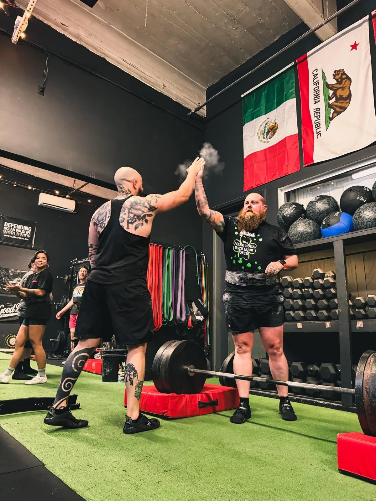 Two men exchange a high five in a gym, with one preparing to lift a barbell while the other congratulates him. The gym has flags of California and the US on the wall, and various workout equipment including kettlebells, resistance bands, and dumbbells.