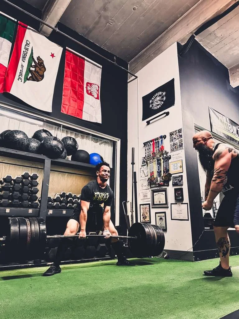 Person performing a squat with a barbell in a gym, featuring weightlifting equipment and green turf area.