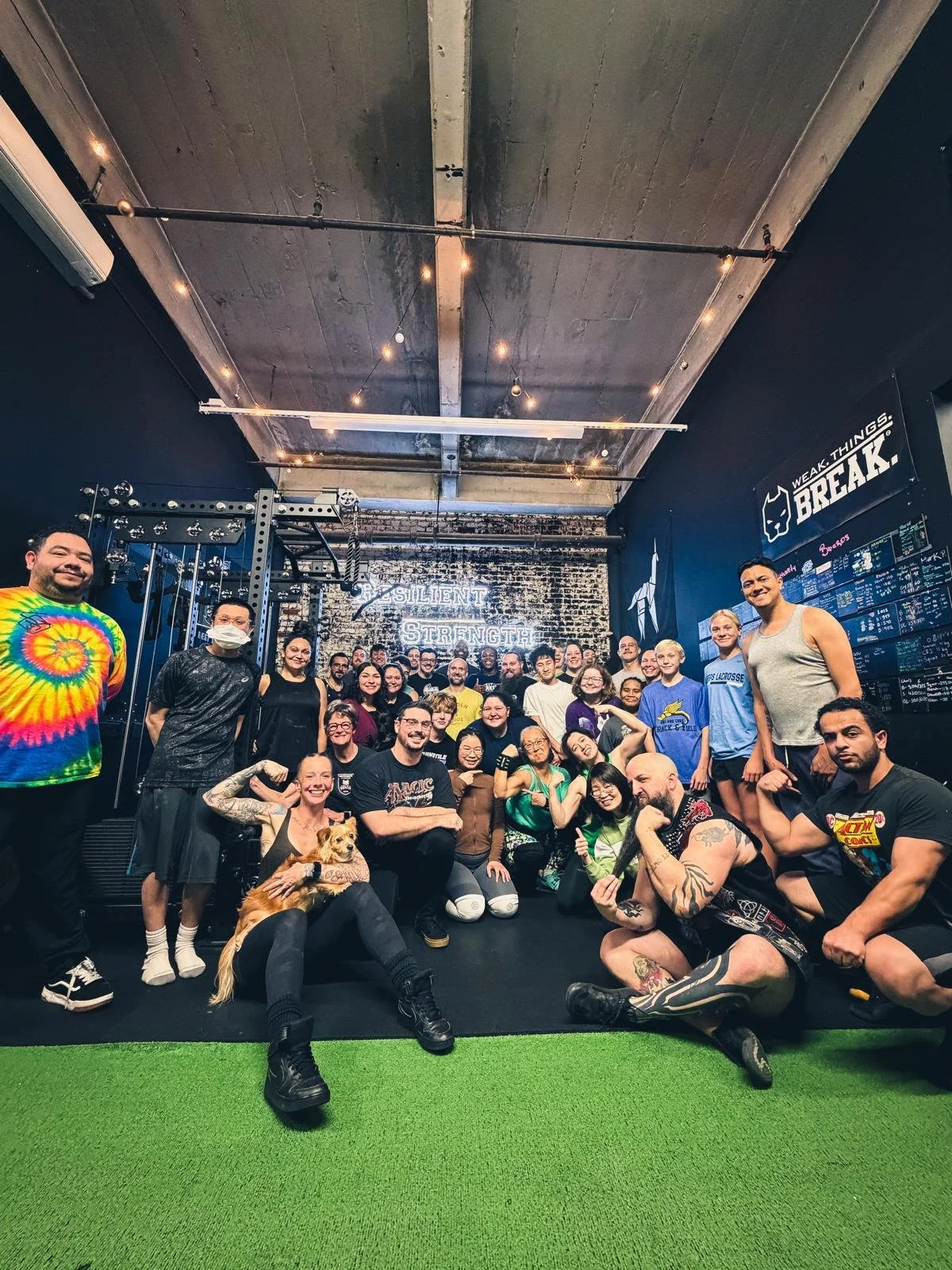 Group of people in a gym, posing for a photo, some smiling and flexing, with a dog in the front. The background features a wall with neon lights reading 'Resilient Strength' and gym equipment.