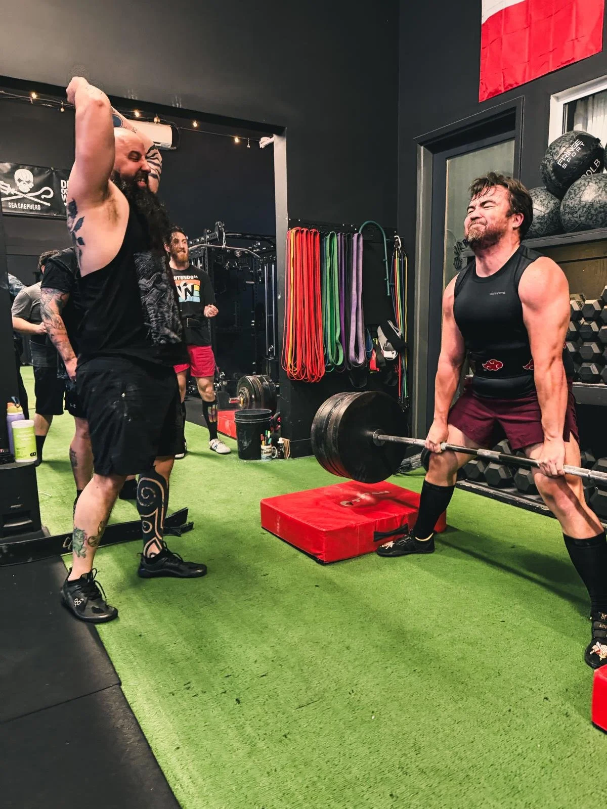 A man performing a deadlift exercise with a heavy barbell in a gym, while others watch and cheer him on, with workout equipment and resistance bands visible in the background.