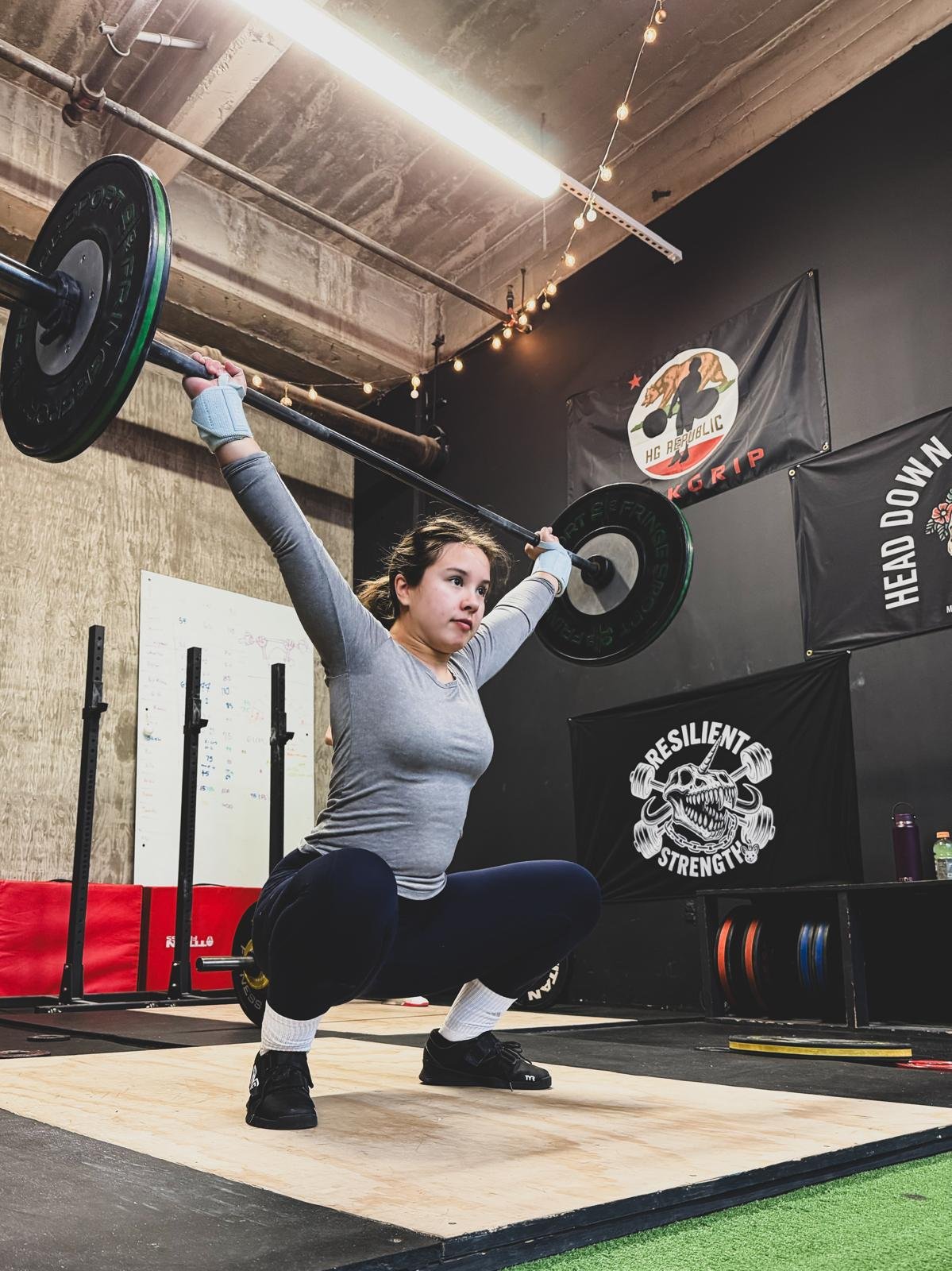 A woman is performing a squat with a barbell overhead in a gym. She is wearing a gray long-sleeve shirt, black pants, and weightlifting shoes. The gym has black walls with posters, including one that says 'Resilient Strength,' and banners for 'KGRIP' and 'Head Down.'