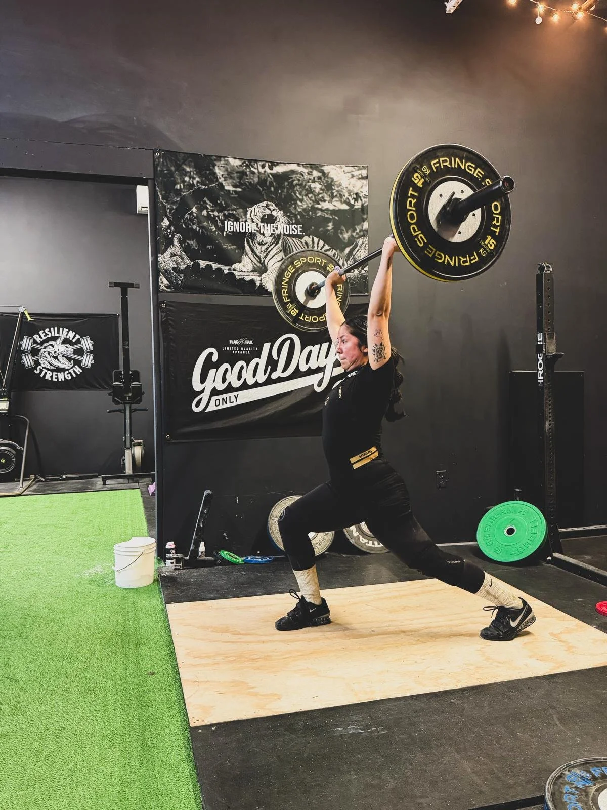 A woman lifting a barbell overhead during weightlifting at a gym, with various weights and gym equipment in the background.