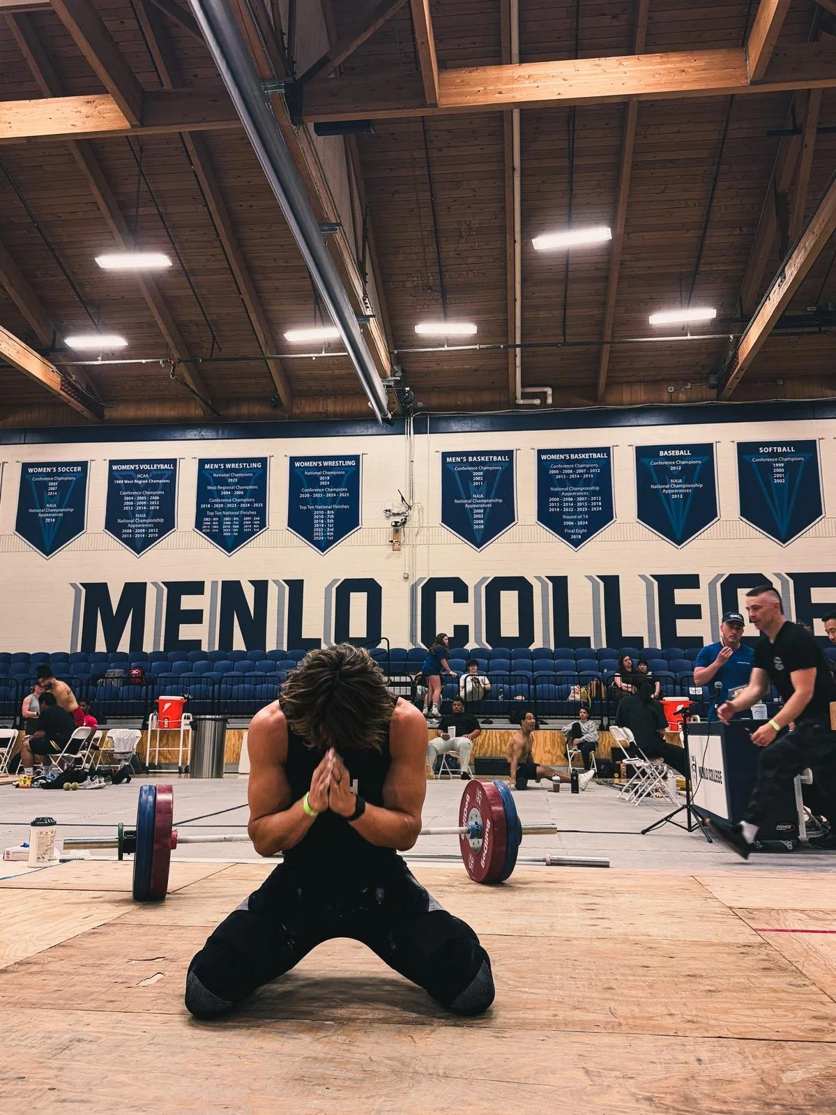 A man kneeling on the floor in front of a barbell with weights, clasping his hands together in a prayer position during a weightlifting competition at Menlo College gymnasium. In the background, multiple athletes and spectators are present, with banners hanging on the wall and a large banner spelling "MENLO COLLEGE."