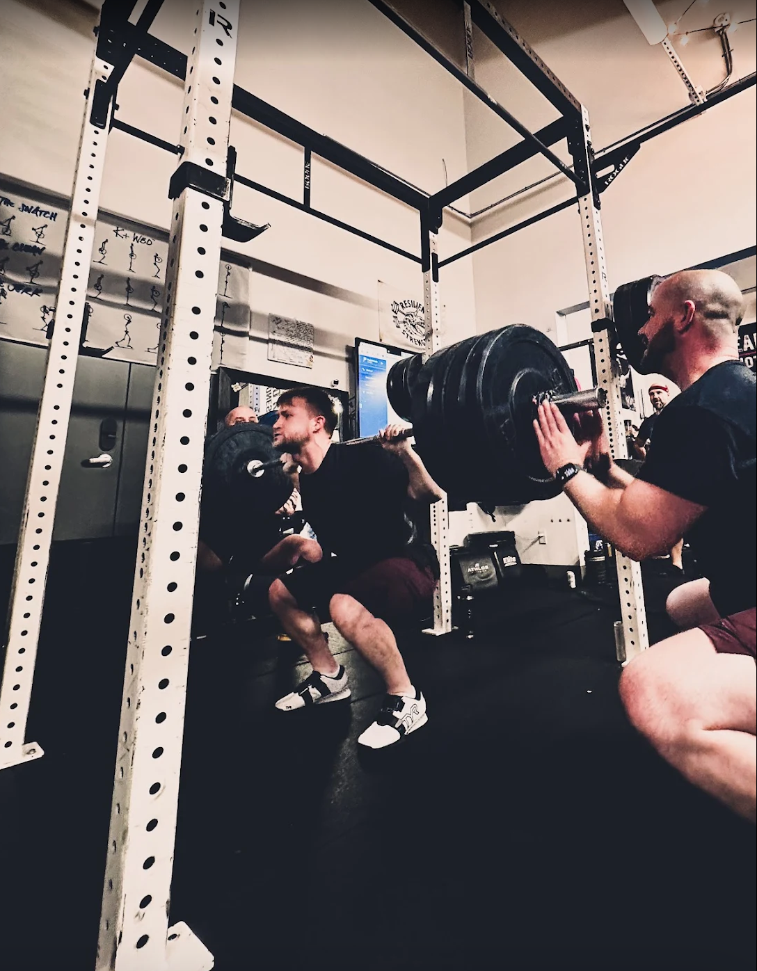 Man squatting and lifting a heavy barbell with weights in an indoor gym, while another man spots him, wearing a mask and holding the barbell.