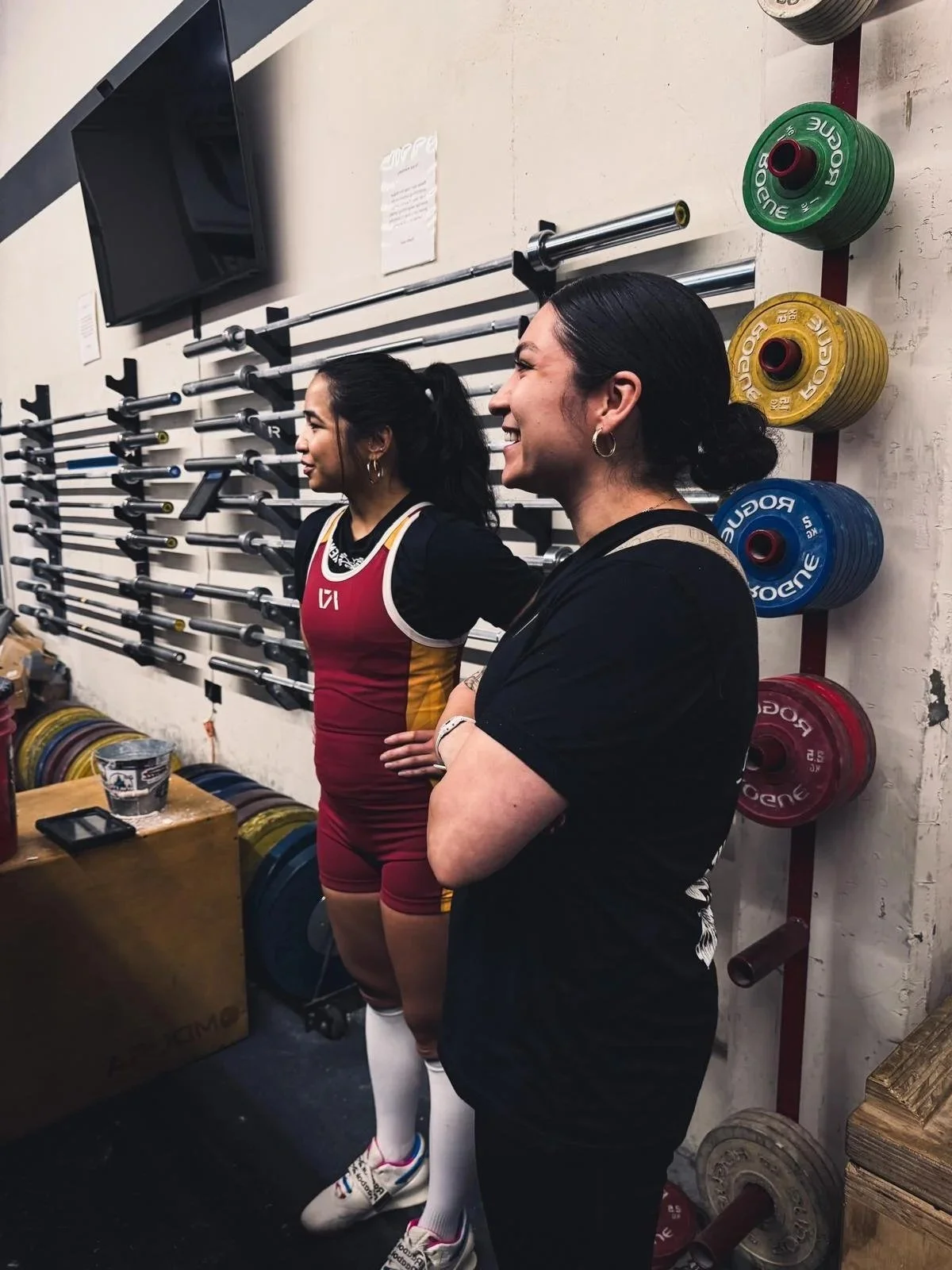 Two women standing in a gym next to a wall with colorful weight plates and weightlifting barbells. One woman is wearing a maroon and gold sports uniform, and the other woman is in a black shirt. Both are smiling and looking to the right.