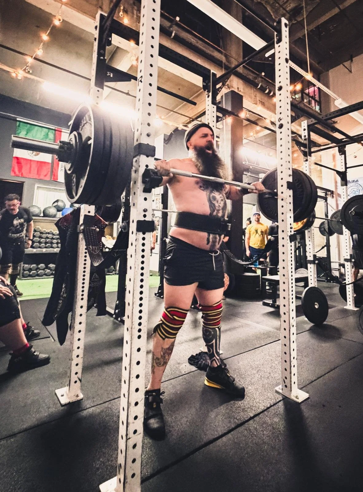 A man with a long beard and tattoos performing a heavy barbell squat in a gym, surrounded by other people and gym equipment.