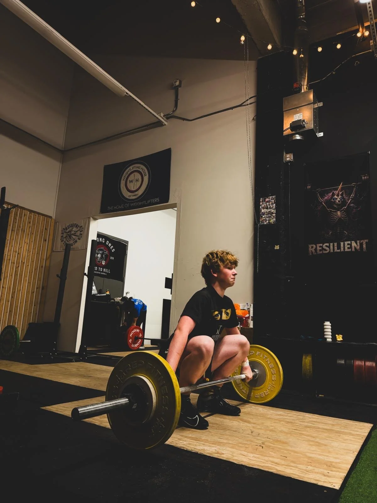 A person performing a weightlifting exercise in a gym, crouching with a barbell loaded with yellow and black weights. The gym has posters on the wall, including one that says "Resilient" and another for a weightlifting house. There is a person in the background lifting weights, and the room is equipped with gym gear and a wooden floor mat.