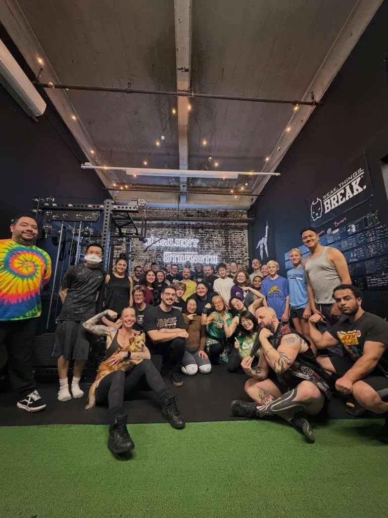 Person performing a barbell squat at a gym, assisted by two spotters, with gym equipment in the background.