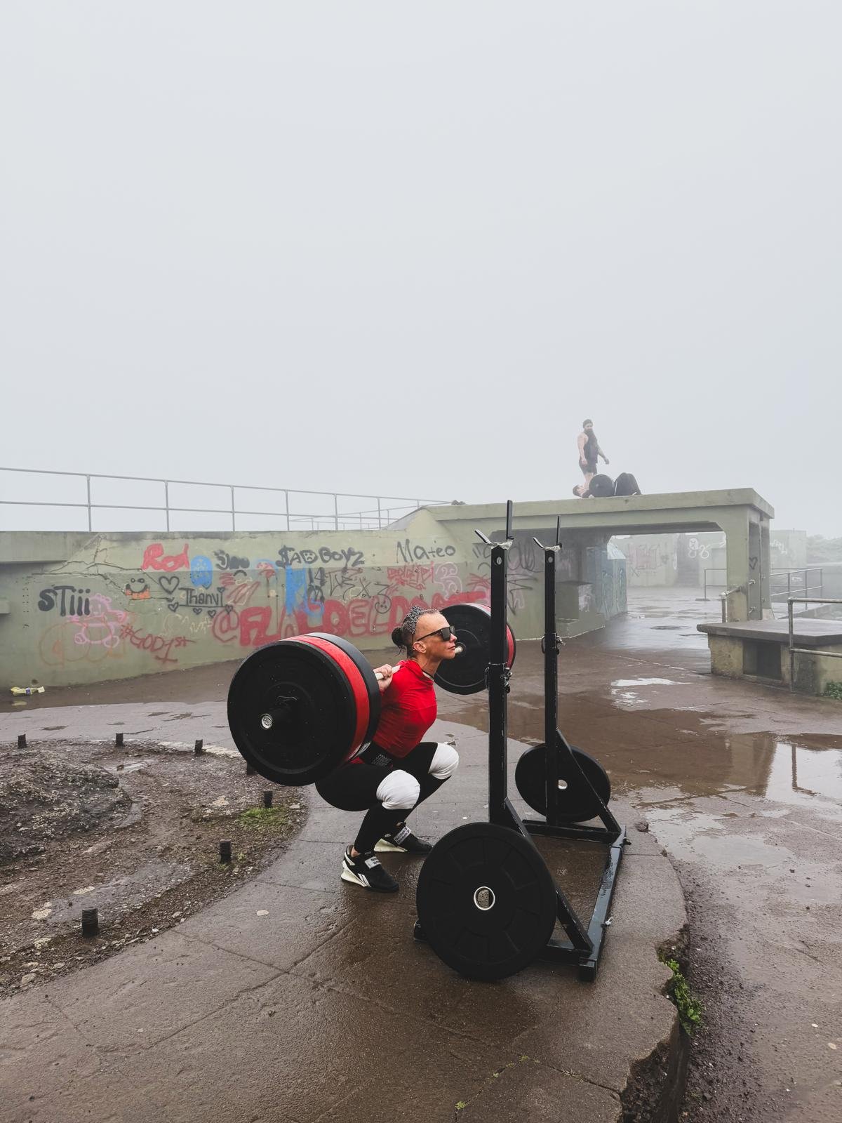 A woman wearing sunglasses and a red shirt squats while lifting a barbell with large weights on a squat rack outdoors. In the background, a foggy setting with graffiti-covered walls and two people standing on a higher platform.